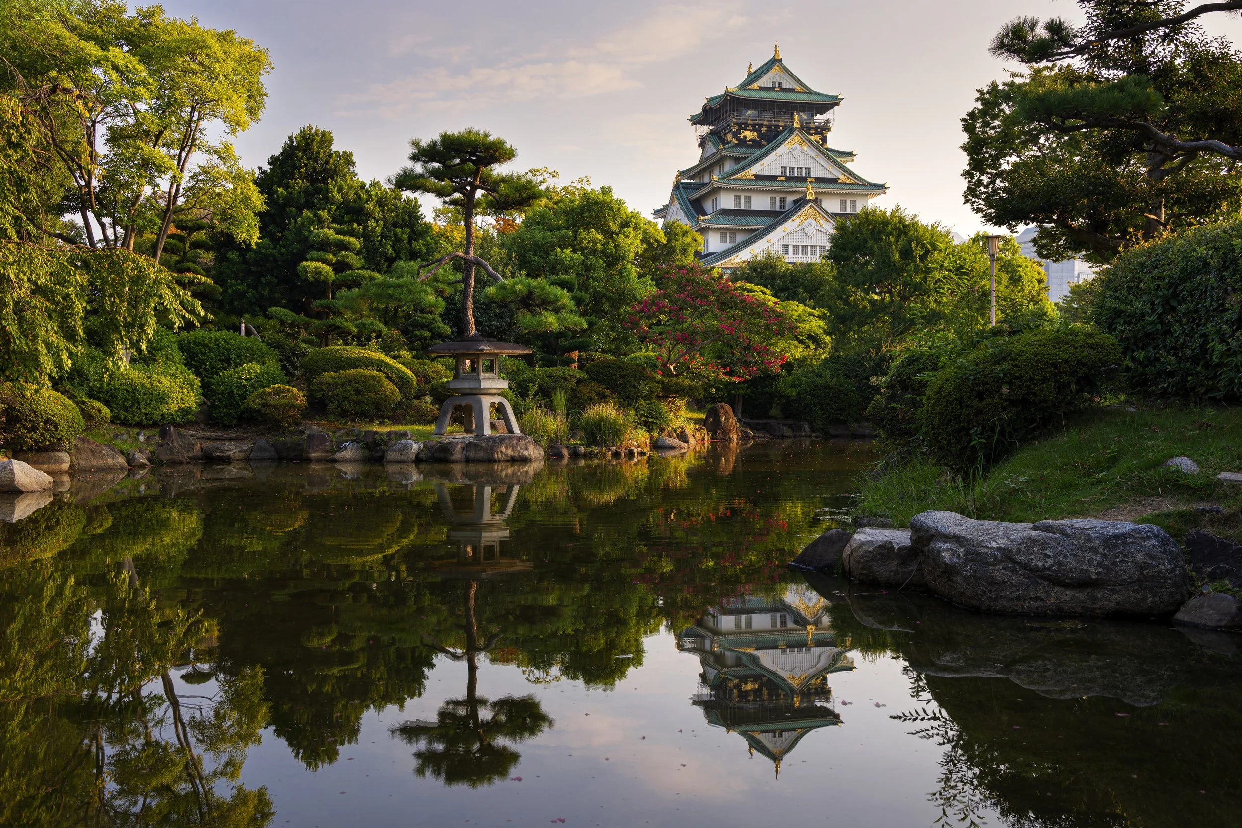 Japanese castle viewed through a lush garden with trees, bushes, rocks, and a pond reflecting the scenery and castle.