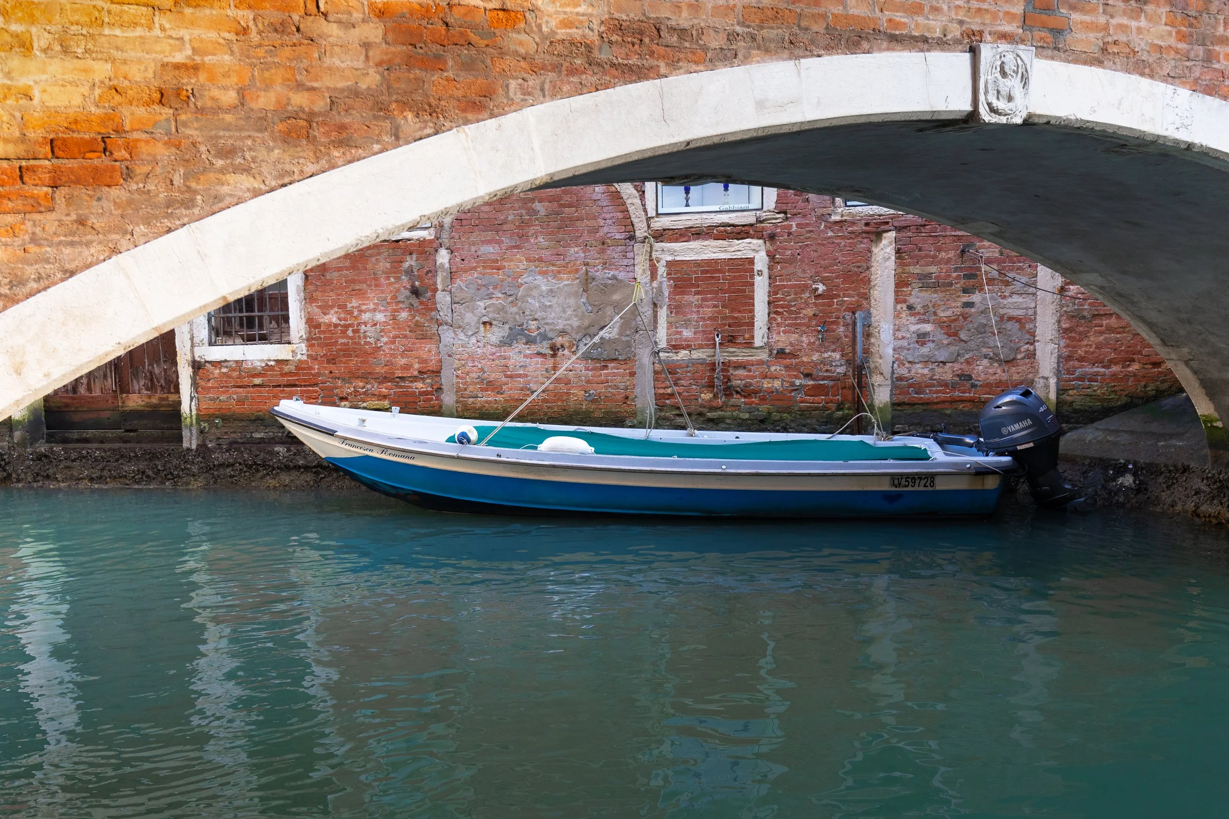 A small boat tied under a brick arch bridge over a canal with brick walls and water.