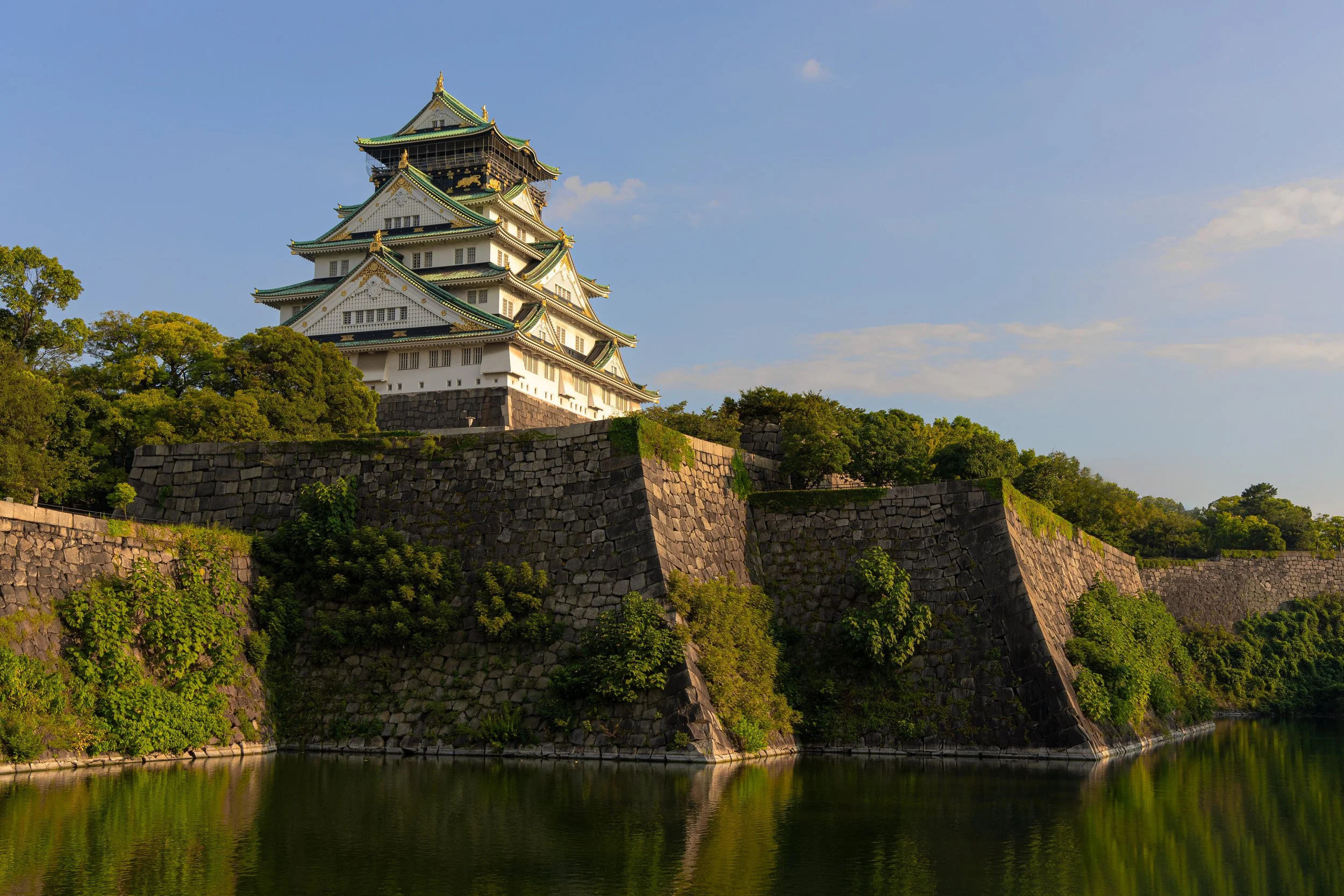 Traditional Japanese castle with white walls and green roof, surrounded by lush greenery and stone fortifications, reflected in a body of water under a partly cloudy sky.