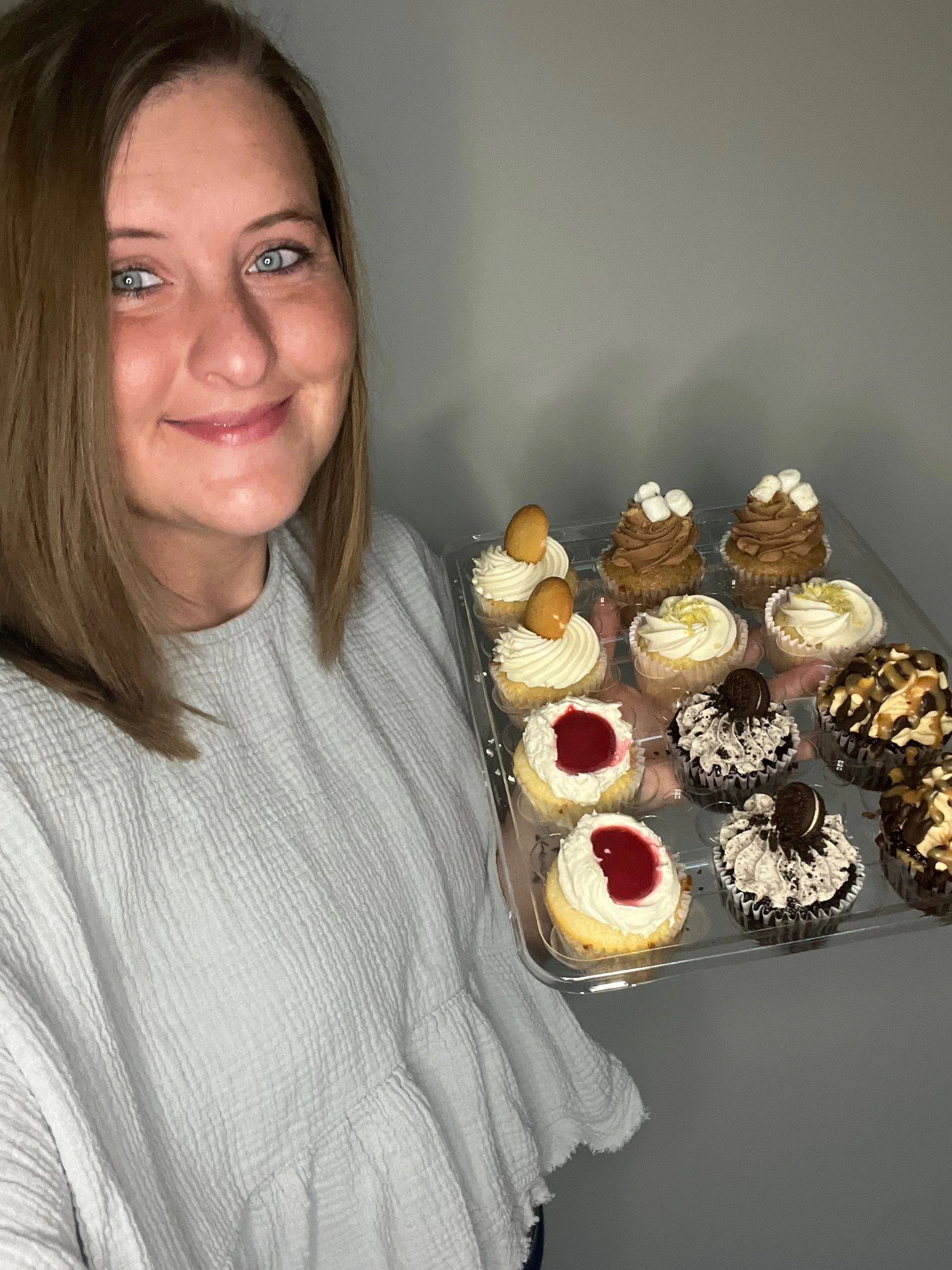 A woman with shoulder-length brown hair and blue eyes smiling while holding a tray of assorted cupcakes.