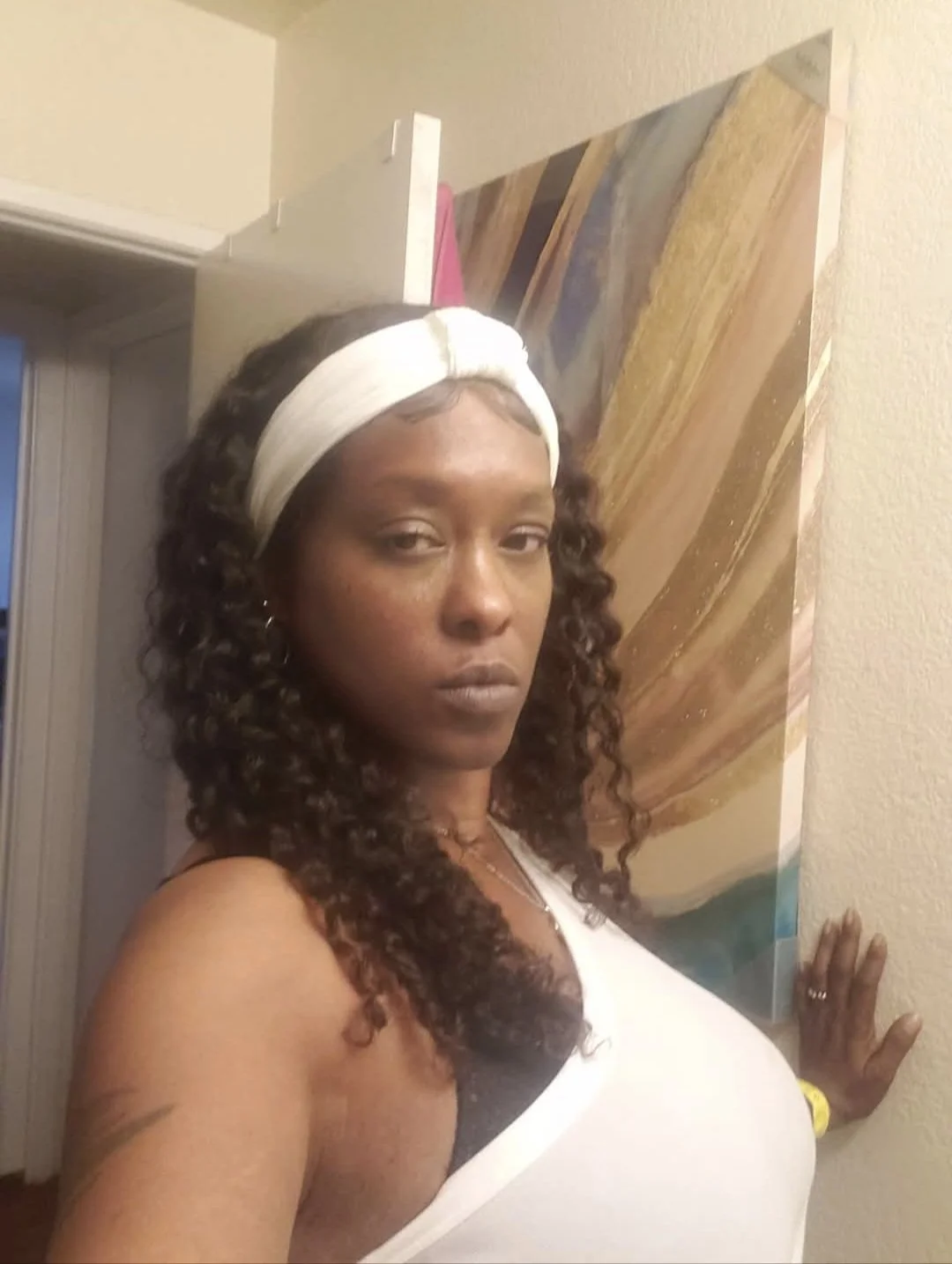 A woman with curly hair wearing a white headband and white tank top standing in front of a colorful abstract painting on a wall.