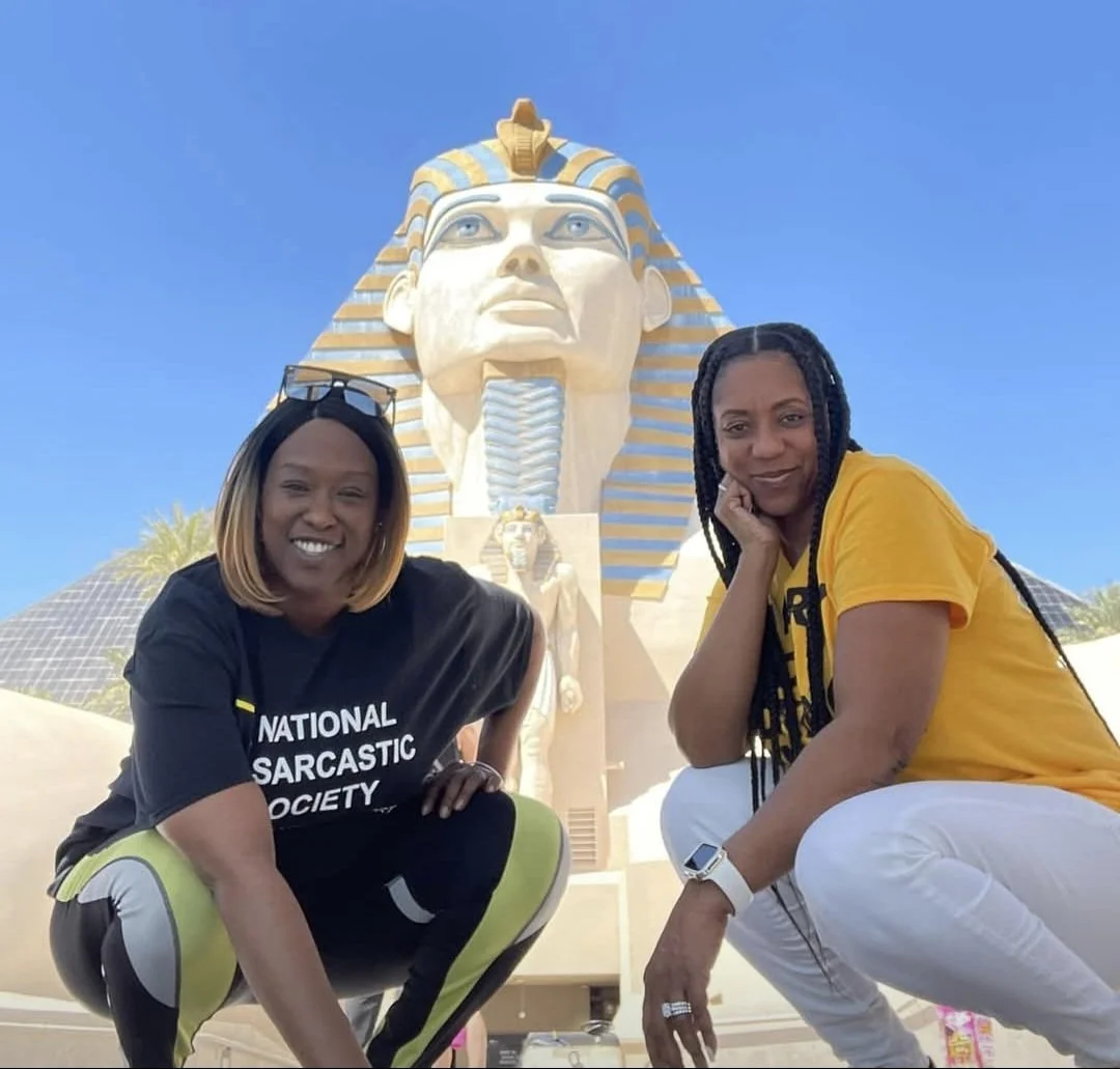 Two women squatting together in front of a giant Egyptian Sphinx statue with a blue sky in the background, one wearing a black t-shirt and the other wearing a yellow t-shirt.