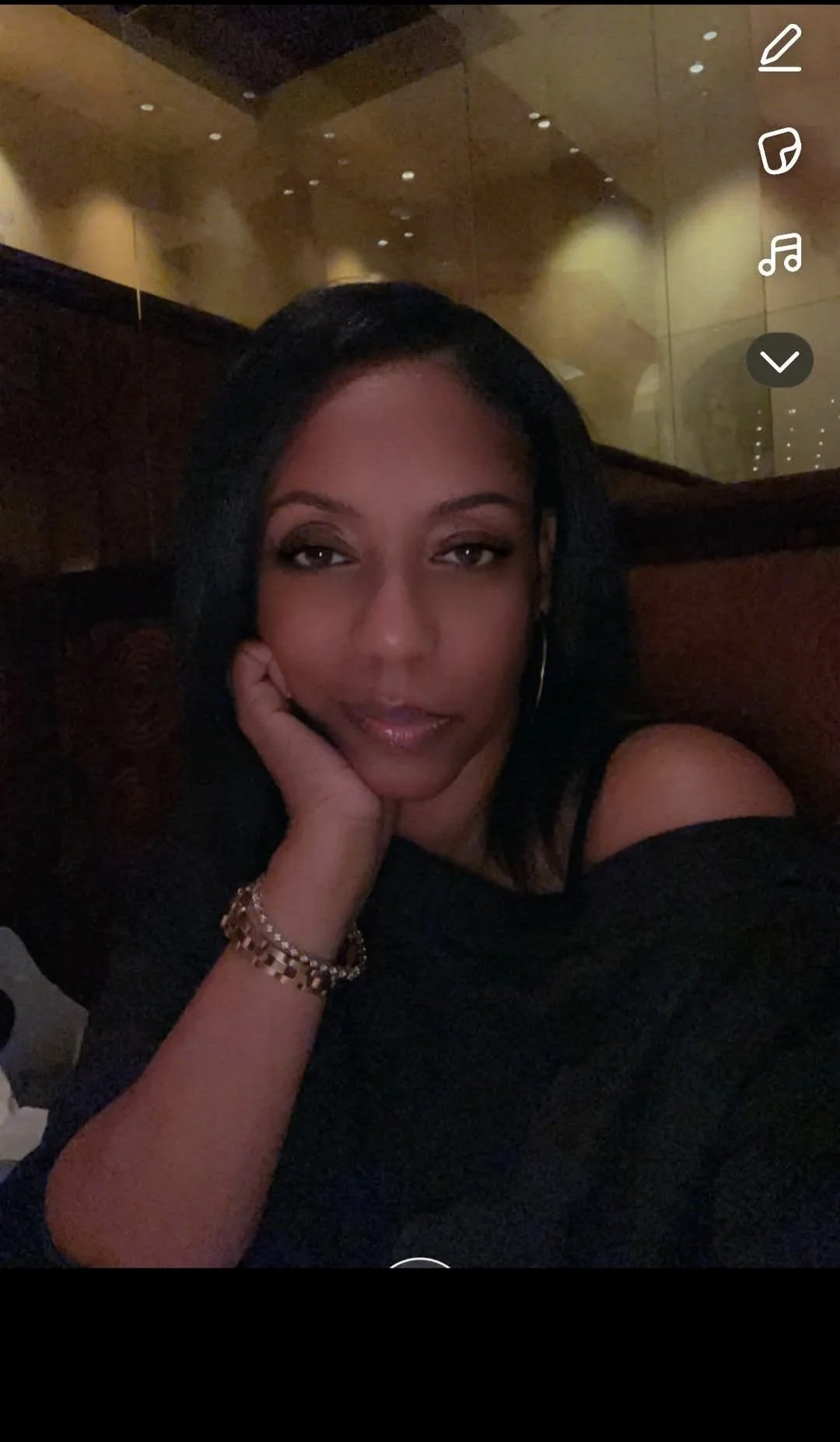 A woman with black hair, wearing makeup and jewelry, resting her face on her hand, sitting in a dimly lit setting.