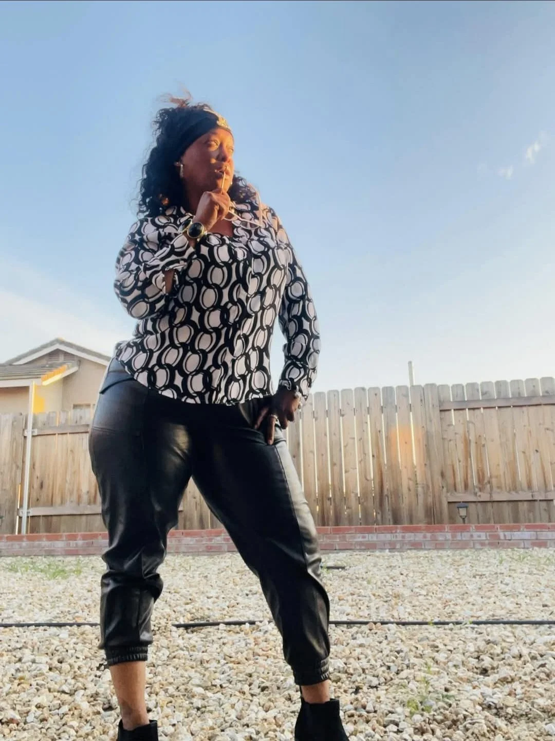 Woman standing outdoors on a gravel surface near a wooden fence, wearing a patterned blouse, leather pants, and a headband with the sun setting in the background.