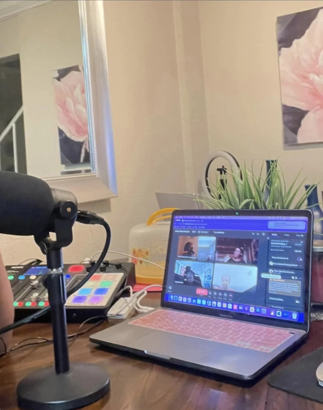 A laptop and audio equipment set up on a wooden table for a virtual meeting or recording, with a potted plant and wall art in the background.