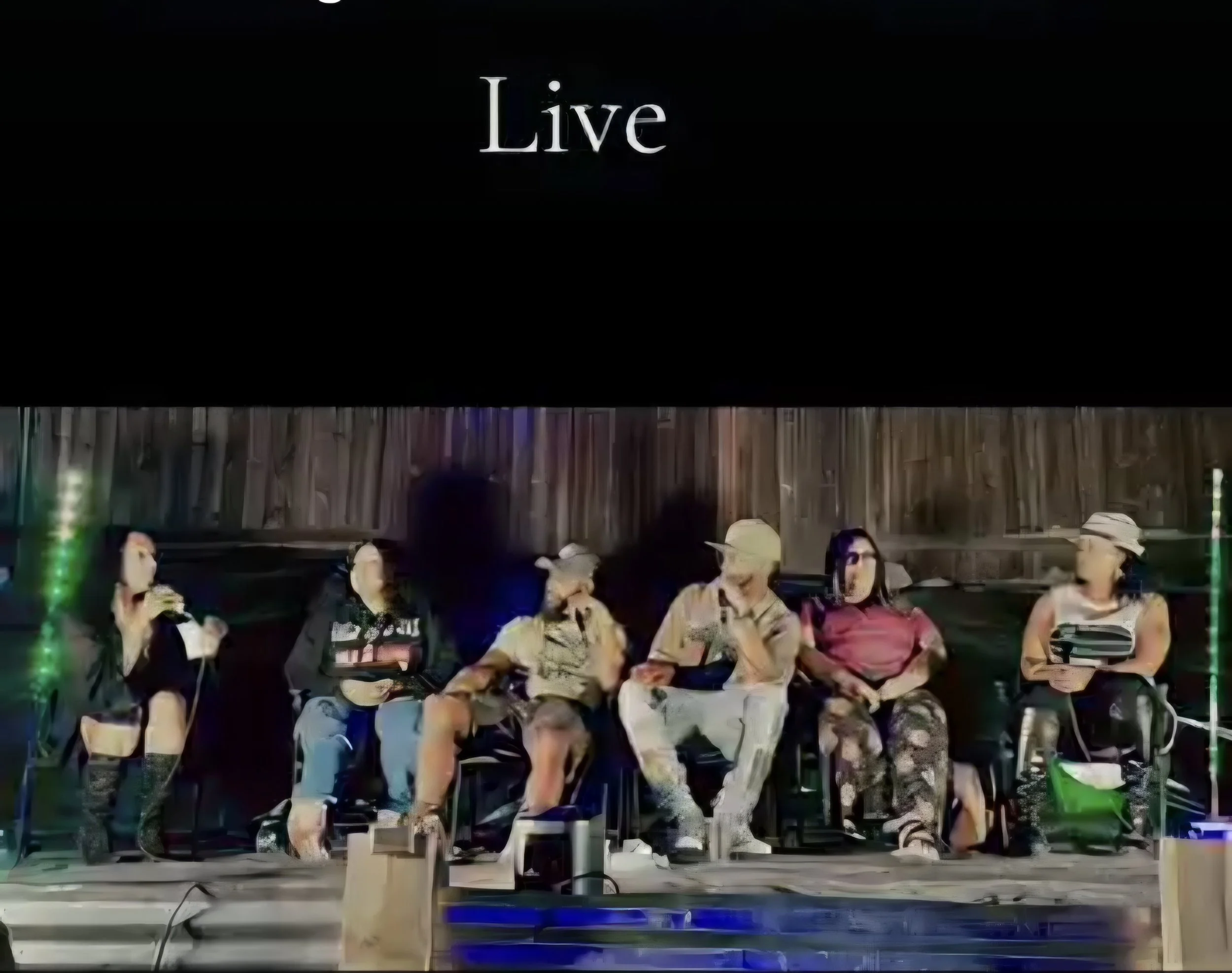 Six women sitting on chairs in a row backstage, preparing for a performance, with a black curtain backdrop and a wooden wall behind them, and the word 'Live' displayed on a screen above them.