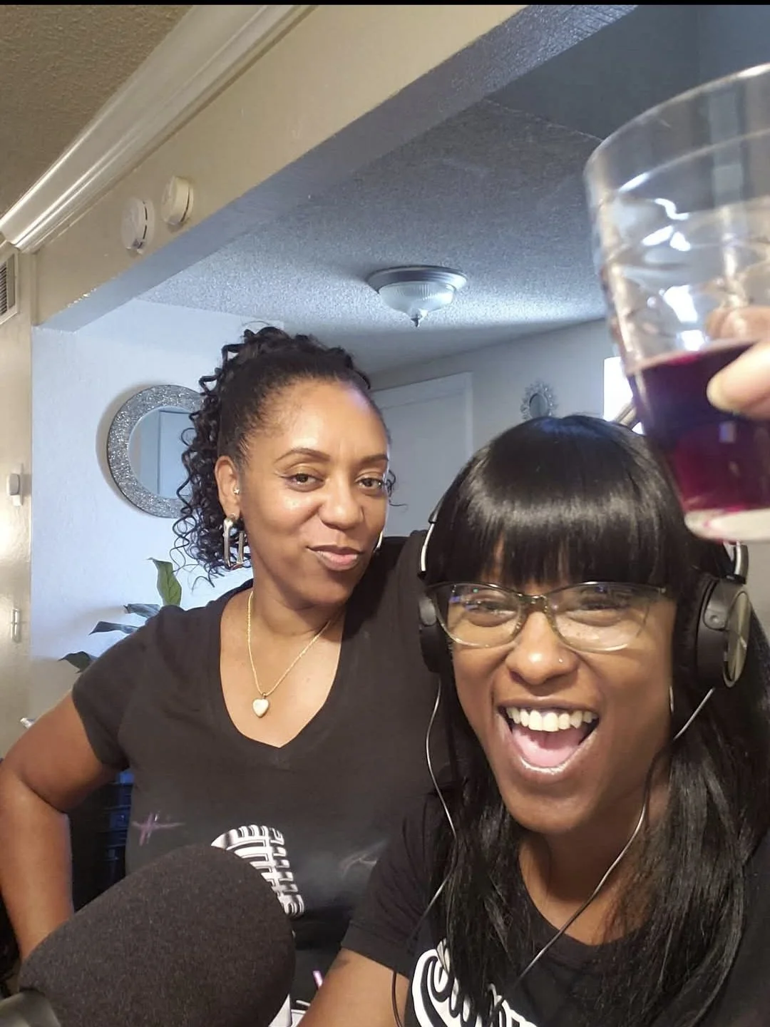 Two women smiling and having a good time. One is holding a glass of red drink and is wearing glasses and headphones. The other has curly hair and is wearing a necklace.