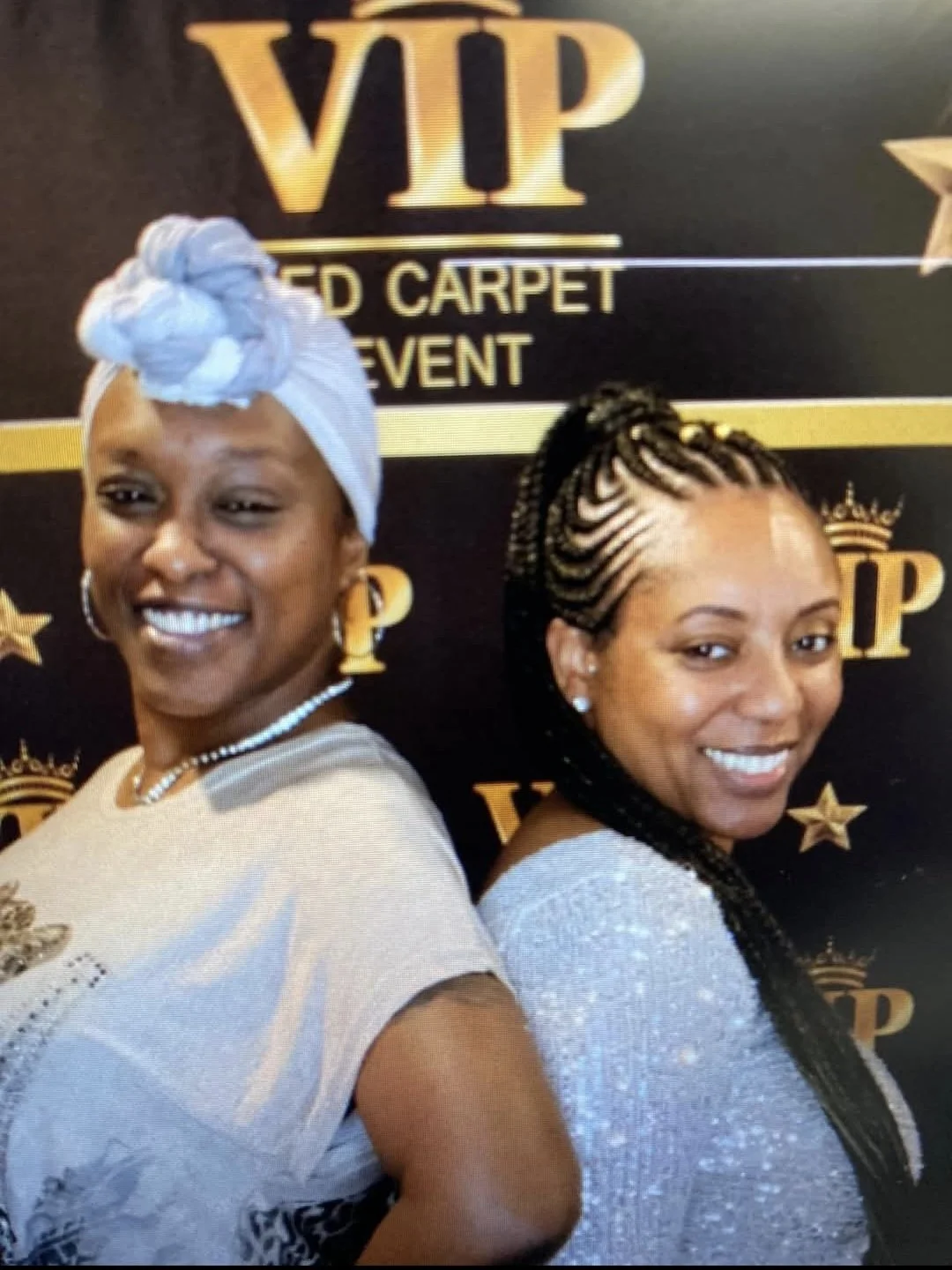 Two women smiling in front of a black backdrop with gold text that reads 'VIP' and 'Red Carpet Event'. One woman is wearing a white and silver outfit, a white headwrap, and gold earrings. The other woman has braided hair with gold beads and is dressed in a silver top.
