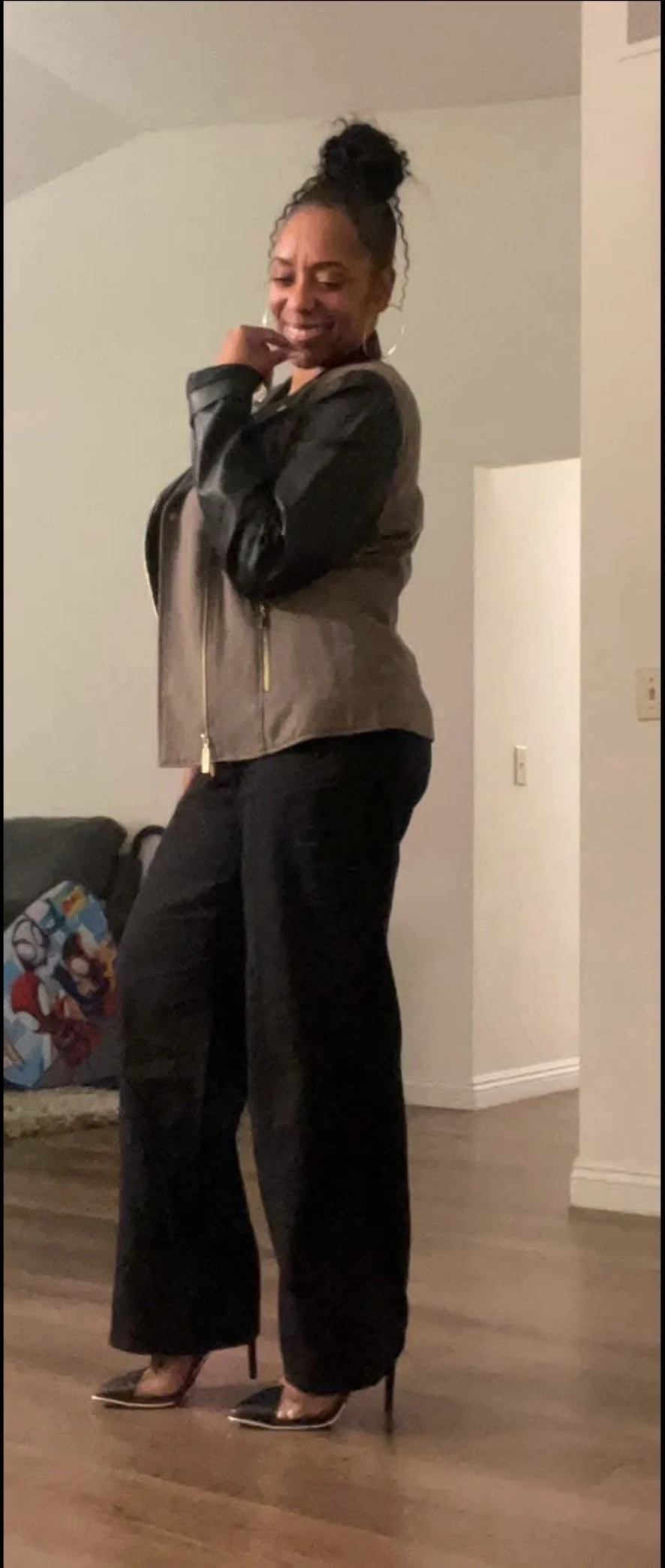 A woman standing in a room, smiling and posing for a photo. She is wearing a black and gray jacket, black pants, and black high-heeled shoes. There is a couch with pillows in the background.