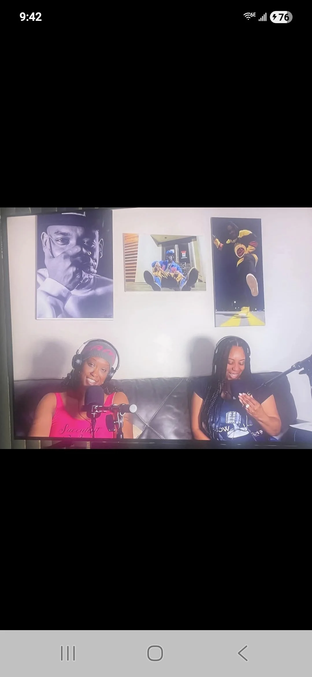 Two women are sitting on a black leather couch, wearing headphones and speaking into microphones. There are three art posters on the wall behind them, featuring portraits and abstract images.