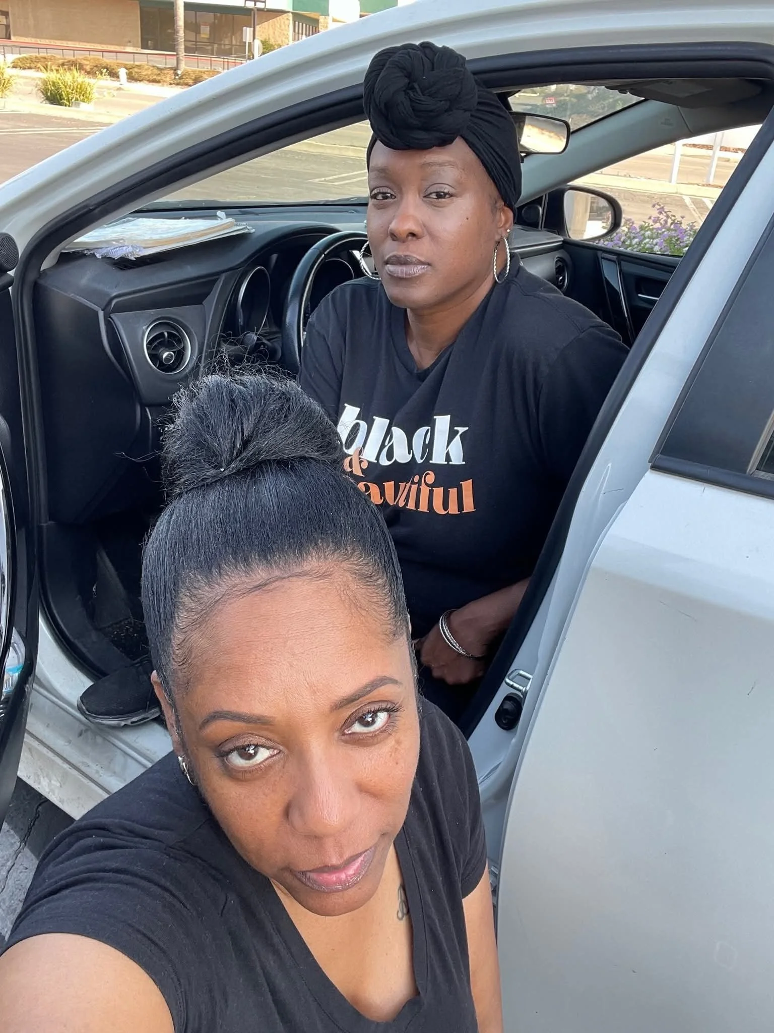 Two women wearing black shirts, one standing outside a white vehicle and the other sitting inside the vehicle, with a parking lot and some buildings in the background.