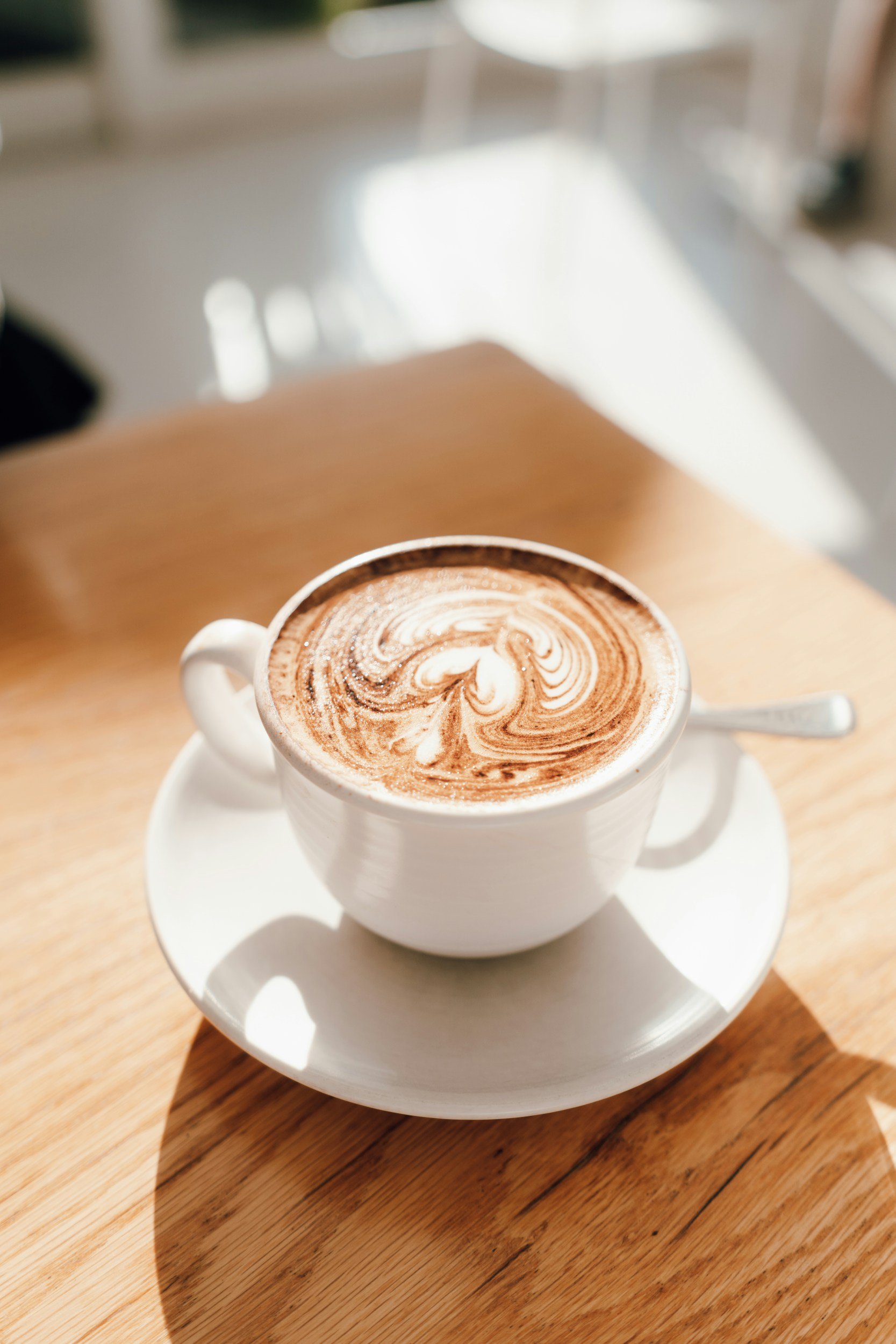 A white ceramic cup of hot chocolate with swirled foam art, placed on a matching saucer and a small spoon, on a wooden table in a brightly lit cafe.