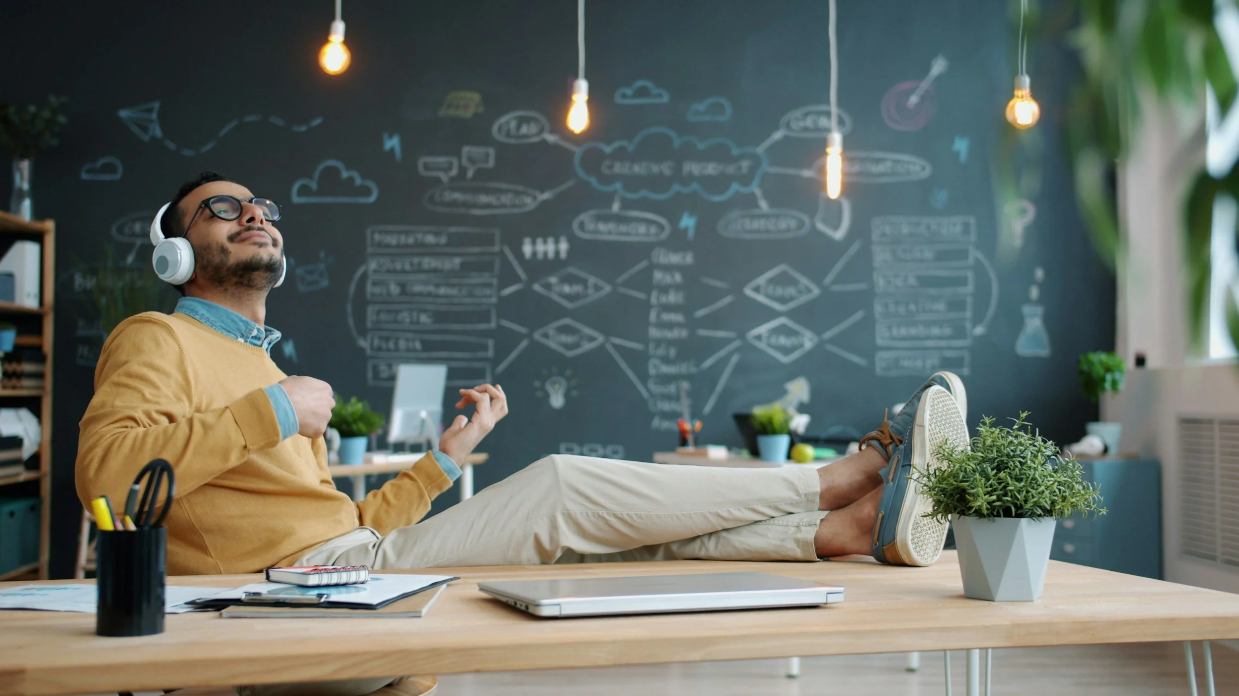 A man relaxing with headphones at a desk in a creative workspace, with a chalkboard wall featuring diagrams and notes in the background.