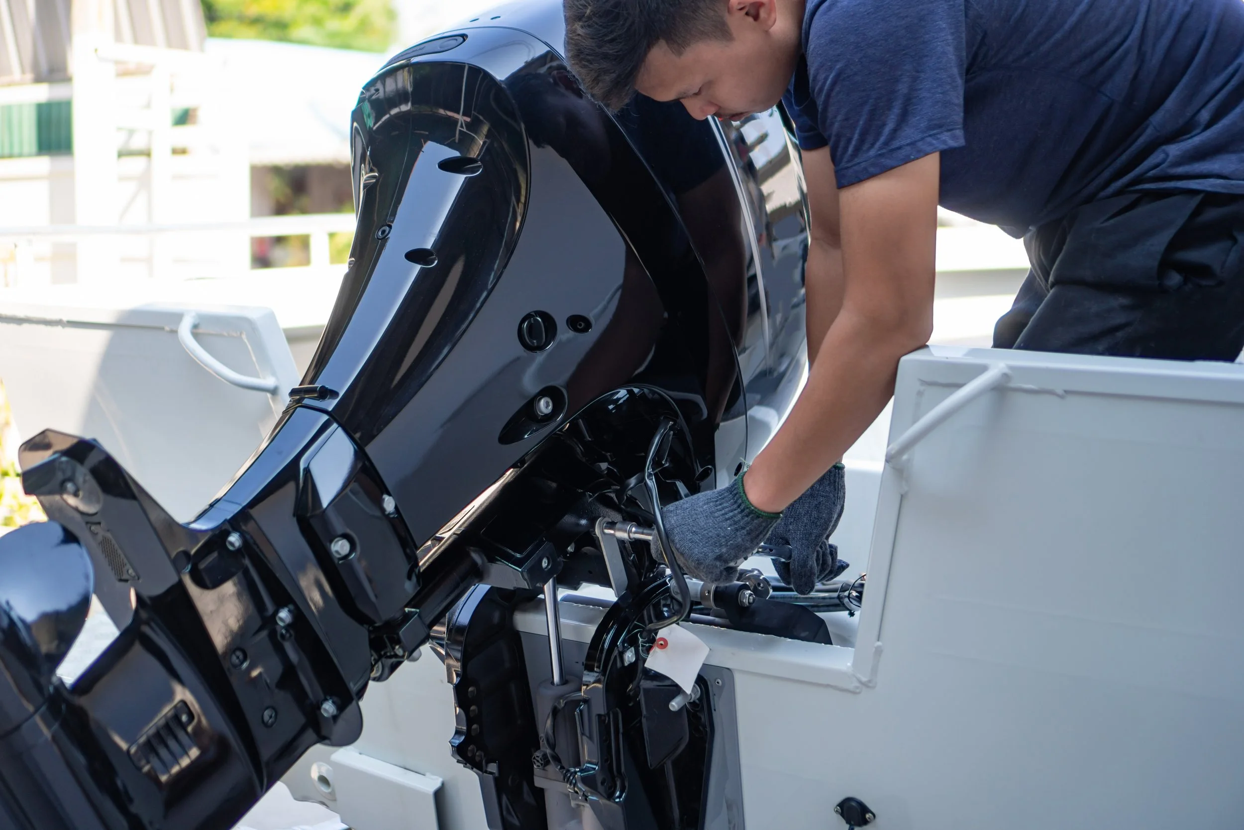A young man working on the engine of a small boat, wearing a blue T-shirt and gray gloves outdoors.