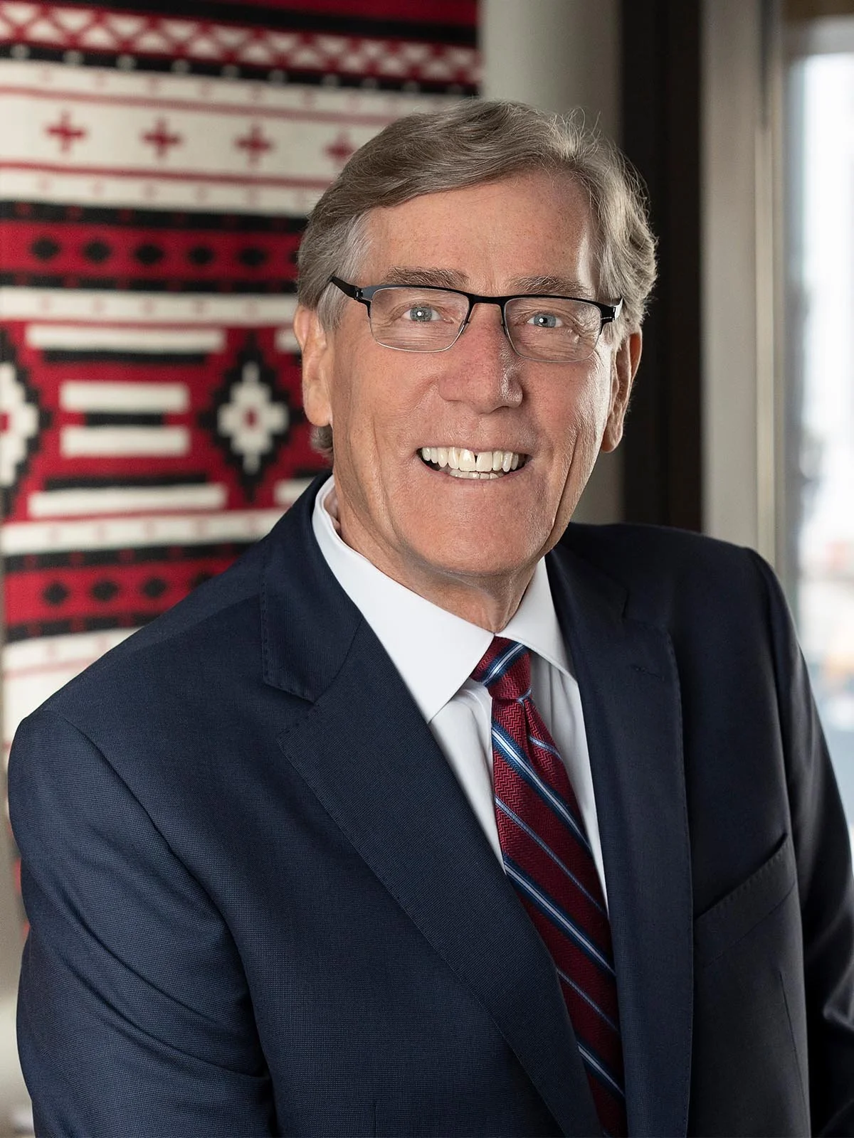 A smiling older man with gray hair and glasses wearing a navy blue suit, white shirt, and a red striped tie, standing indoors near a window with a patterned textile hanging behind him.