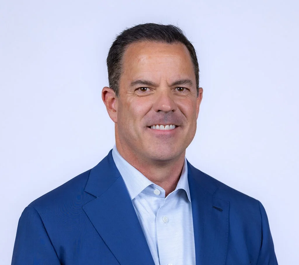 Professional headshot of a man in a blue suit and light blue shirt smiling against a plain background.