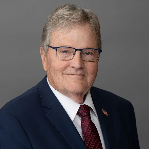 Portrait of an older man with gray hair and glasses, wearing a dark suit, white shirt, and red patterned tie, with a small American flag pin on his lapel, against a gray background.