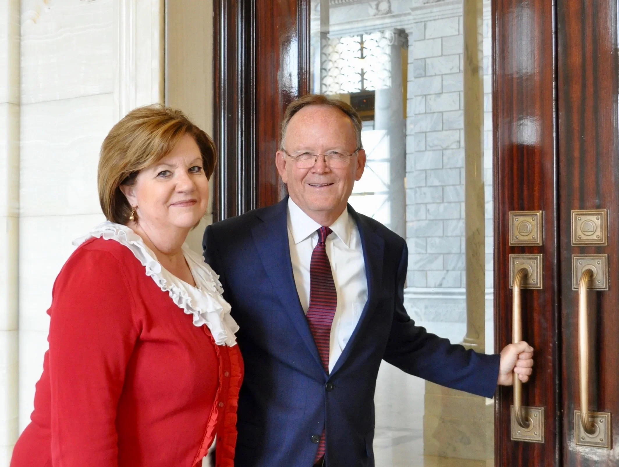 Two people, a woman in a red dress with white ruffled collar and a man in a dark suit with a striped tie, standing at an open doorway with brass handles, smiling at the camera.