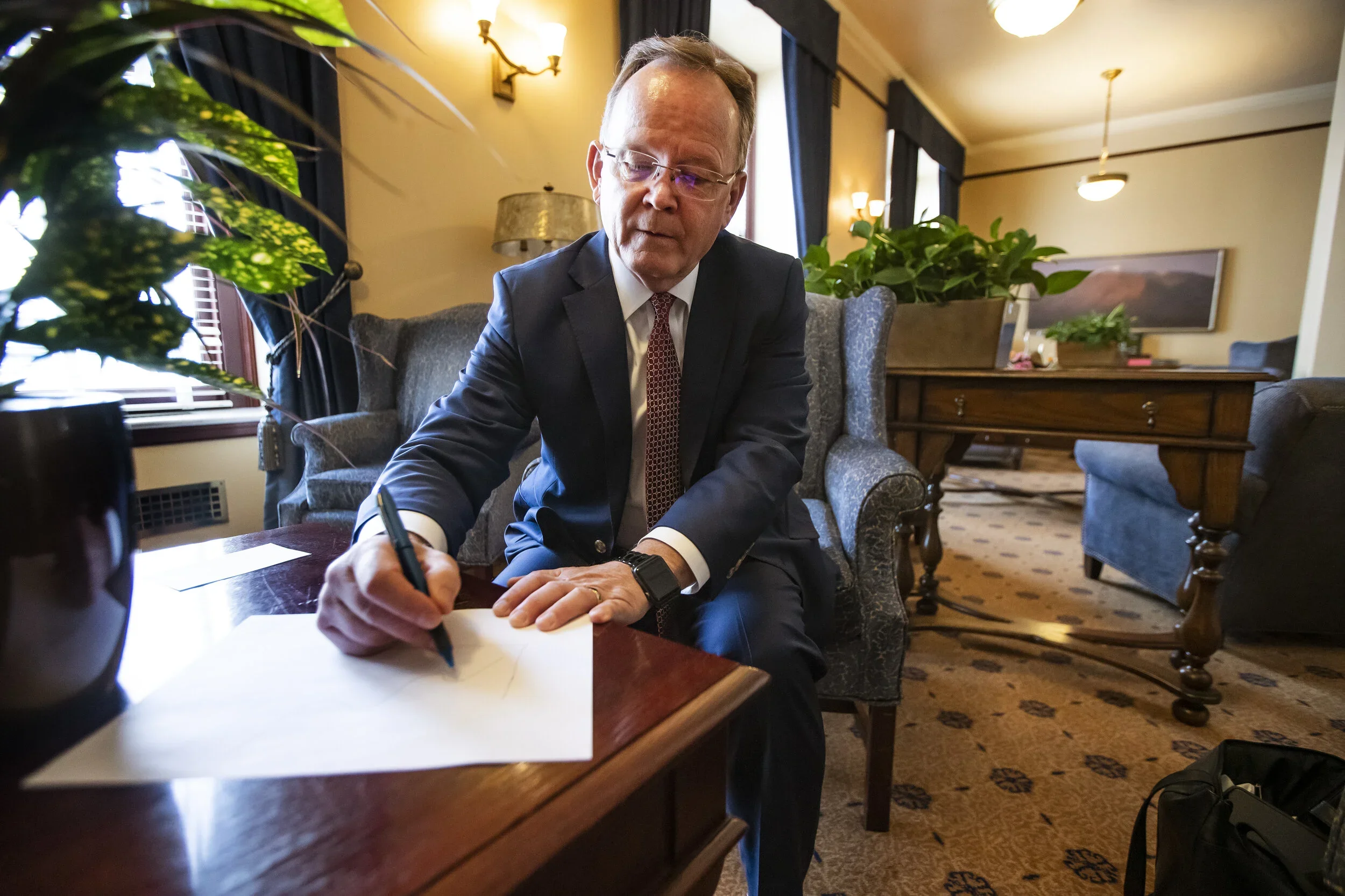 A man in a dark suit and red tie is sitting at a wooden table, writing on a piece of paper with a black pen. The background features a cozy, well-lit room with a sofa, potted plants, and framed artwork on the walls.