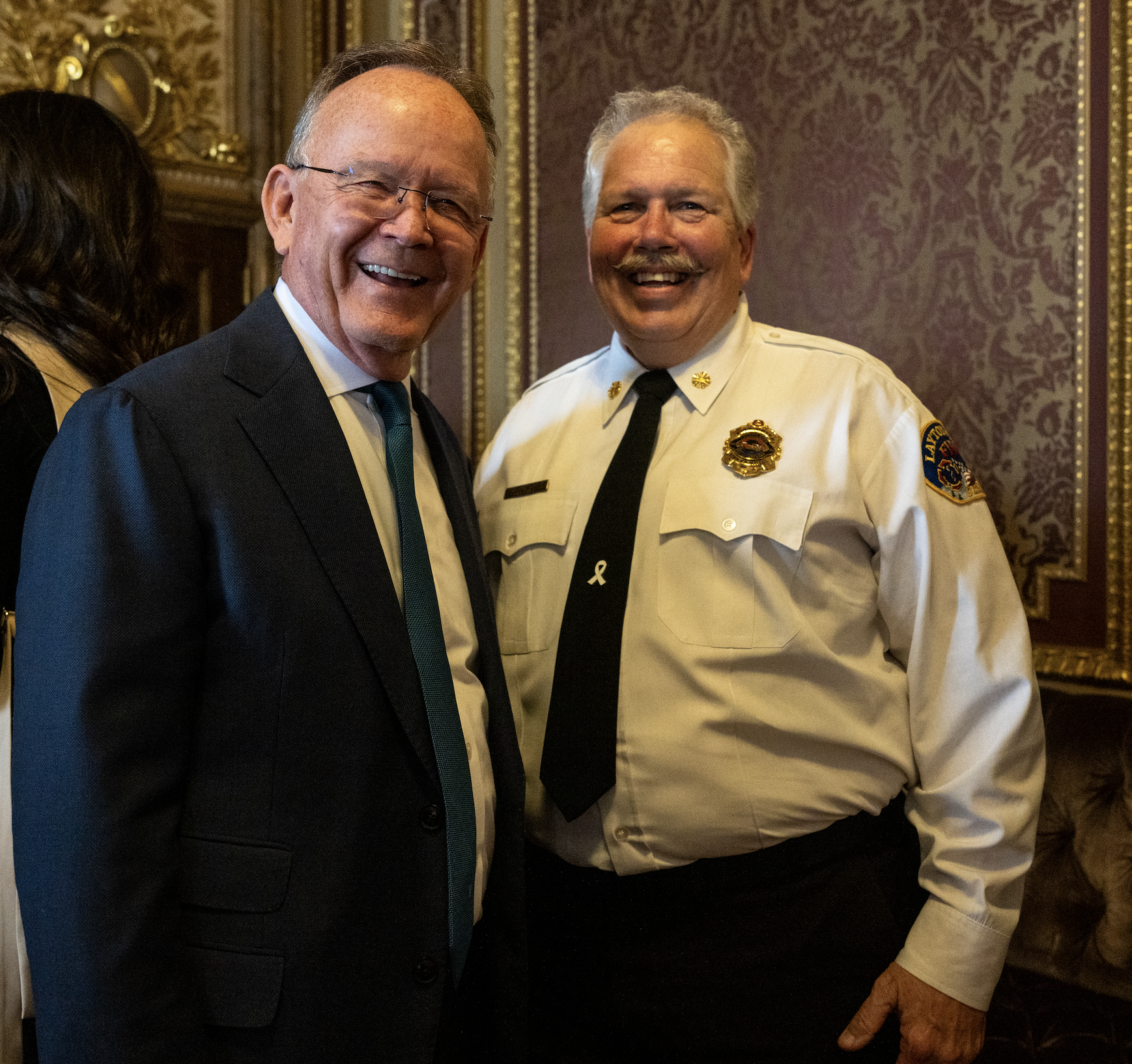 Two smiling men, one in a black suit and striped tie, the other in a white uniform shirt with a badge, pins, and a patch on the sleeve, standing in a richly decorated room with ornate wallpaper and gold accents.