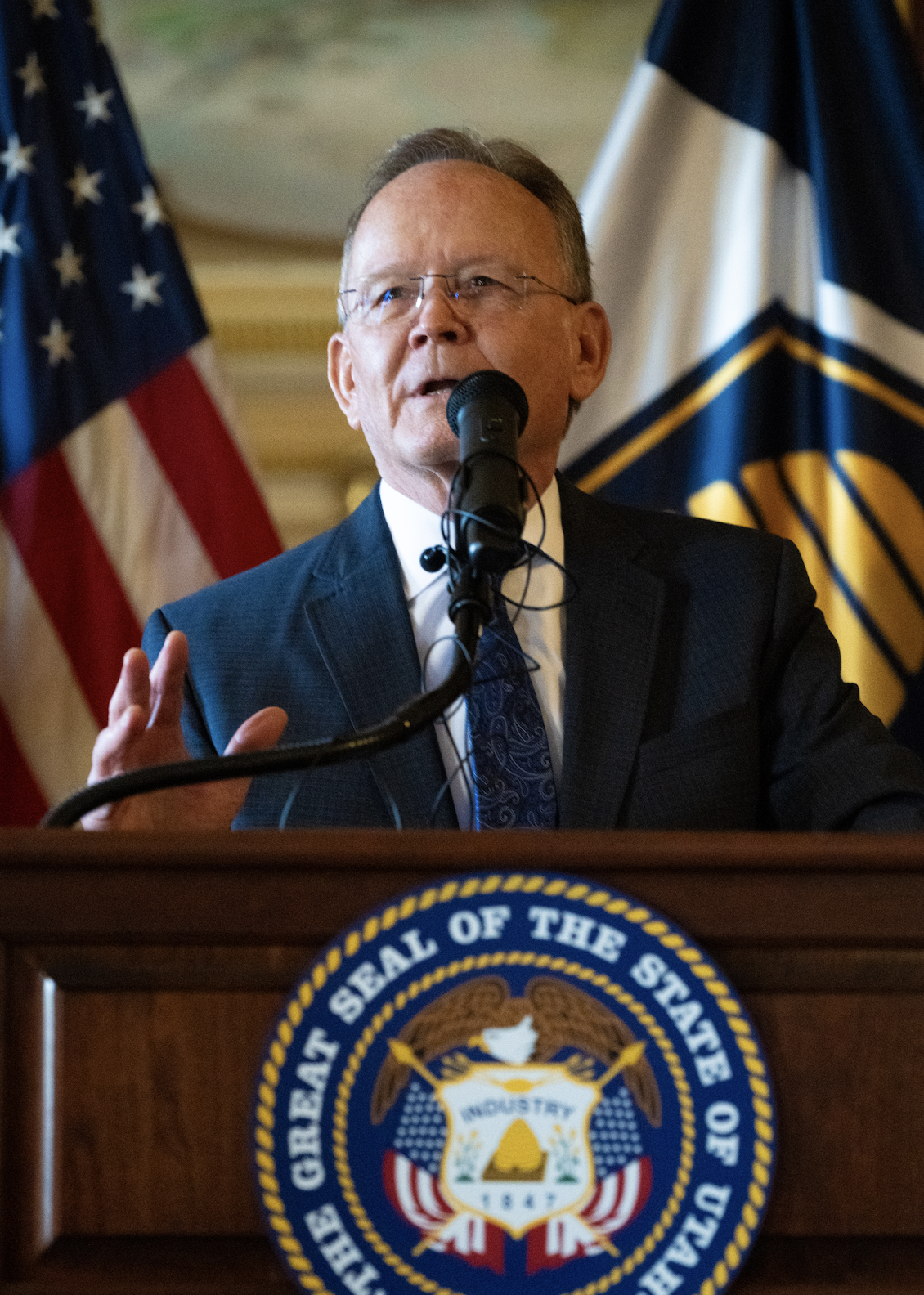 A man in a suit and glasses speaking at a podium with a microphone. The podium has a seal that reads 'The Great Seal of the State of Maine'. In the background, there are two flags, one American and one which appears to be a state flag.
