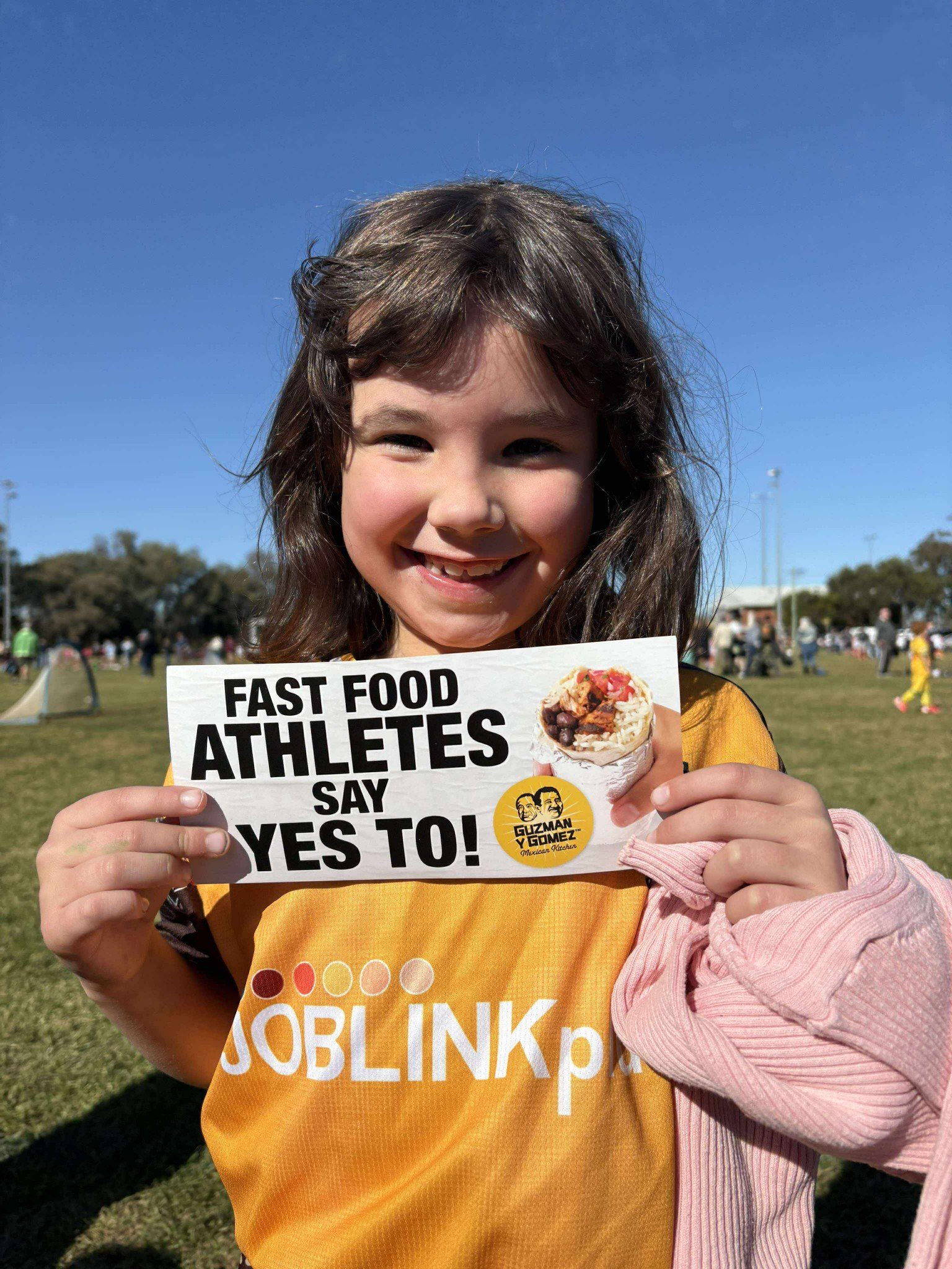 A young girl smiling and holding a sign that says "Fast Food Athletes Say Yes To!" with an image of a taco and a logo for Guzman y Gomez Mexican Kitchen, outdoors on a sunny day in a grassy park with other people in the background.