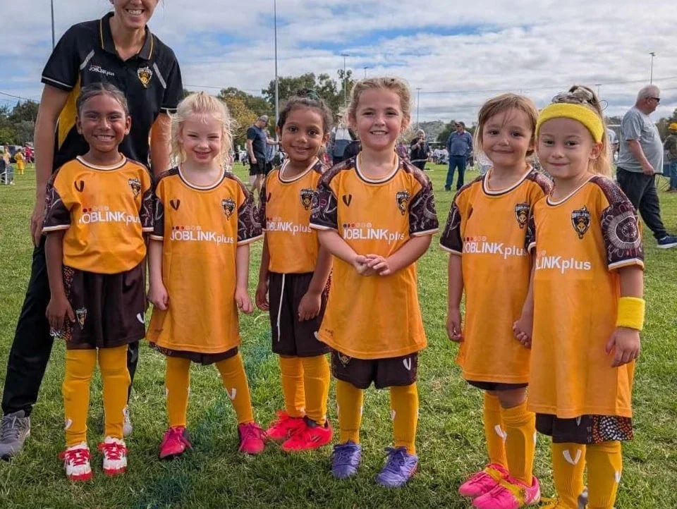 Group of young girls in yellow and black soccer uniforms with a coach, standing on a grassy field at a sports event.
