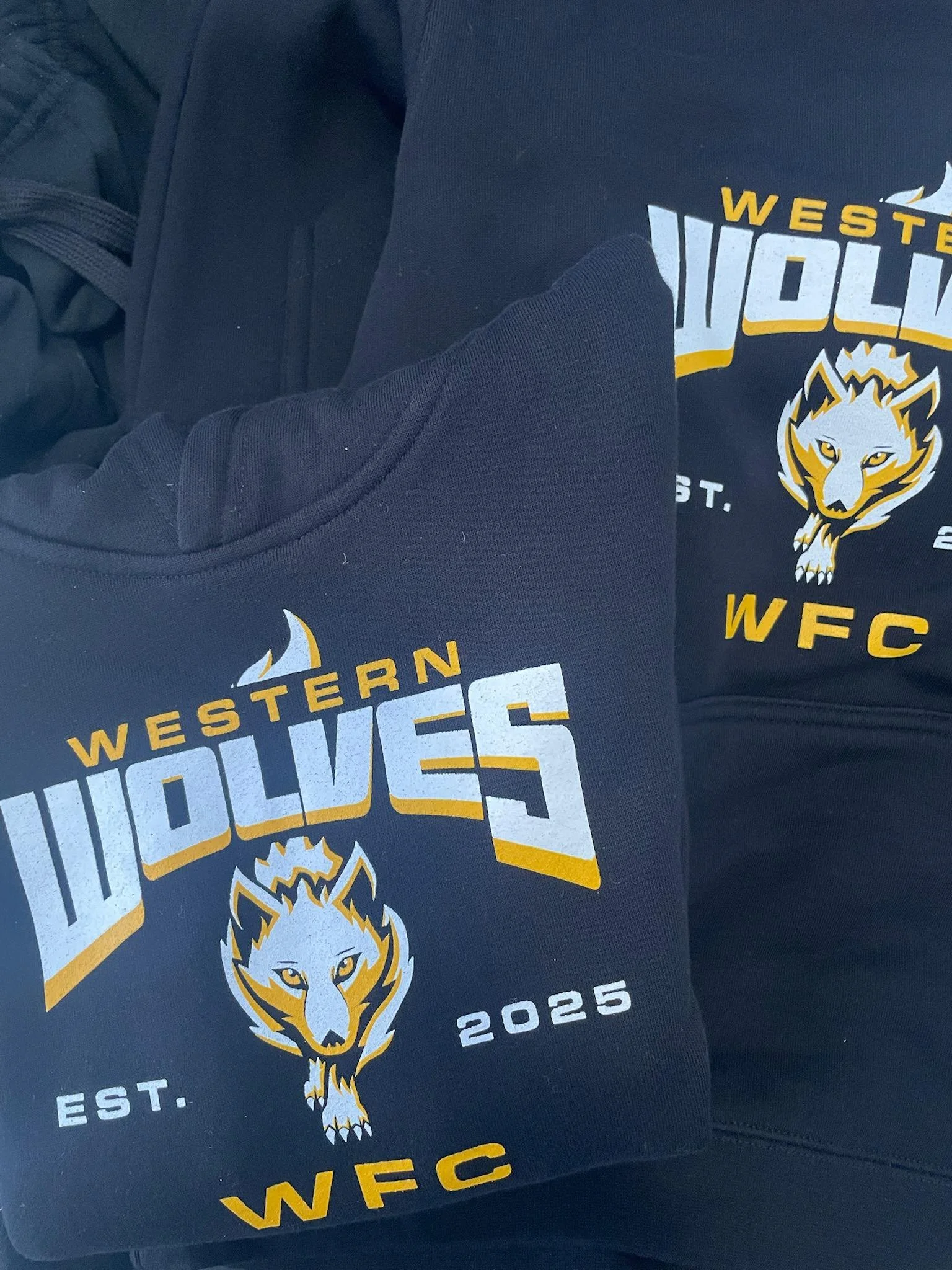 Two black sweatshirts with a logo for Western Wolves sports team, featuring a wolf's head and text 'Western Wolves,' 'WFC,' and 'EST. 2025'.