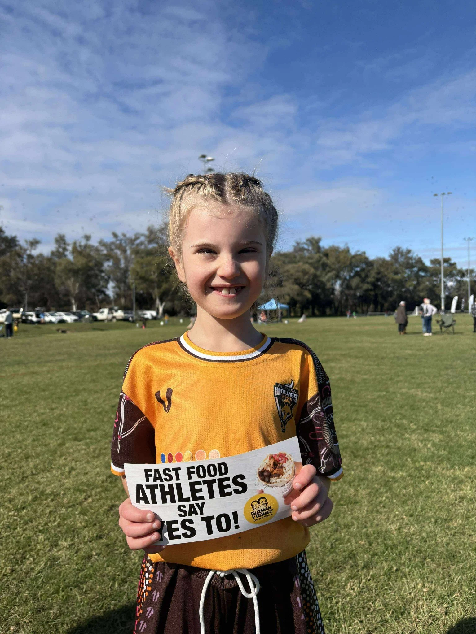 A young girl with blonde hair in braids, smiling and holding a sign that says "Fast Food Athletes Say Yes To!" at an outdoor sports field on a sunny day.
