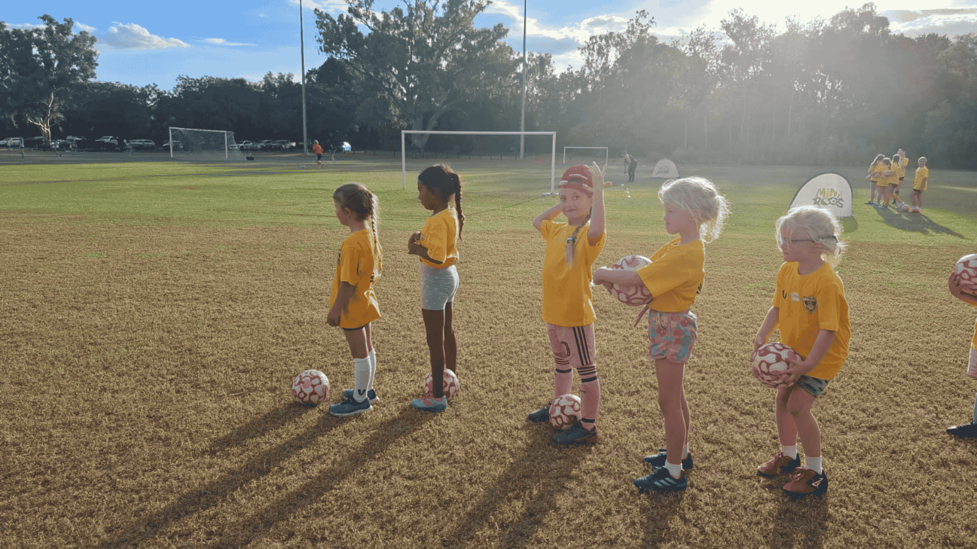 Six young girls in yellow jerseys standing in a line on a grassy field, each holding a soccer ball, preparing for a soccer practice or game in late afternoon with long shadows and trees in the background.