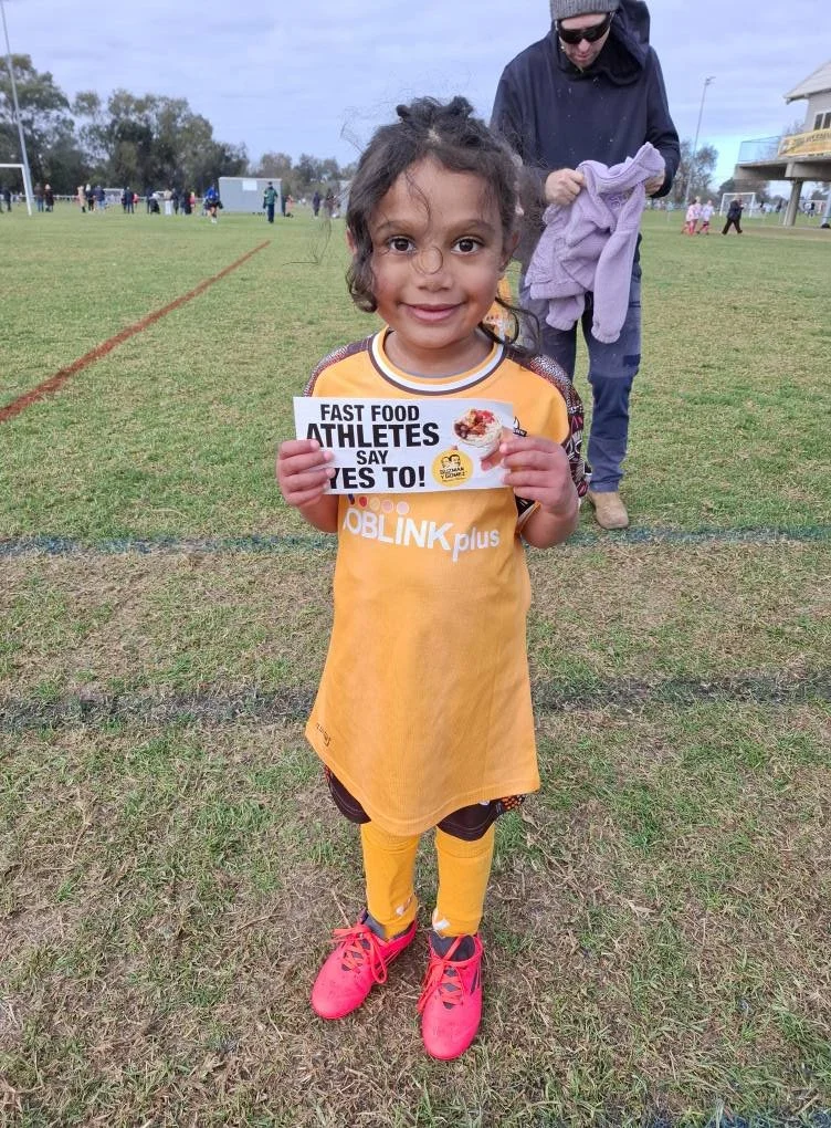 Young girl wearing a yellow sports uniform and pink shoes, holding a sign promoting fast food athletes, standing on a sports field with people in the background.