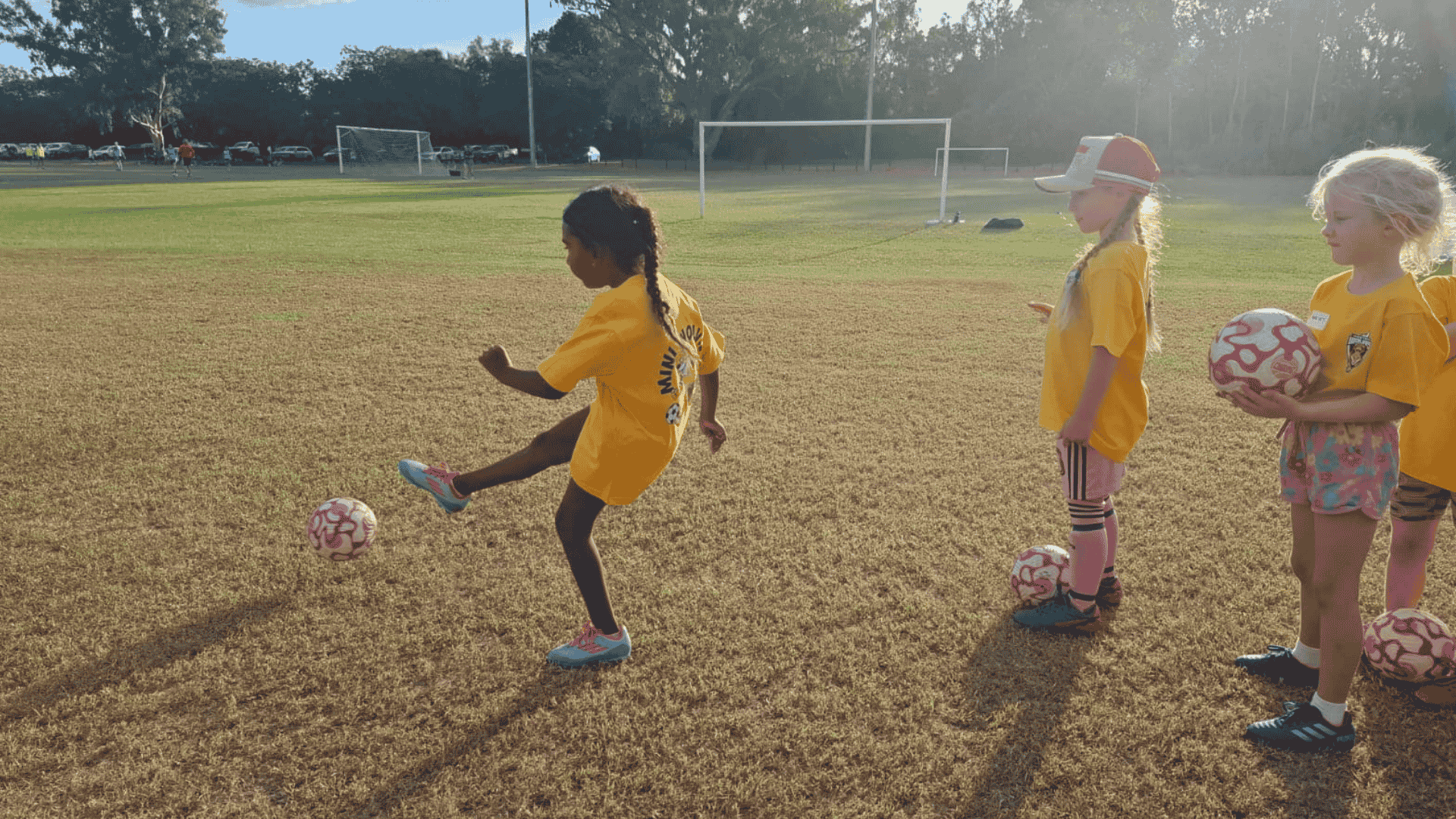 Children in yellow shirts training on a soccer field with pink and white soccer balls, with goalposts and parked cars in the background.