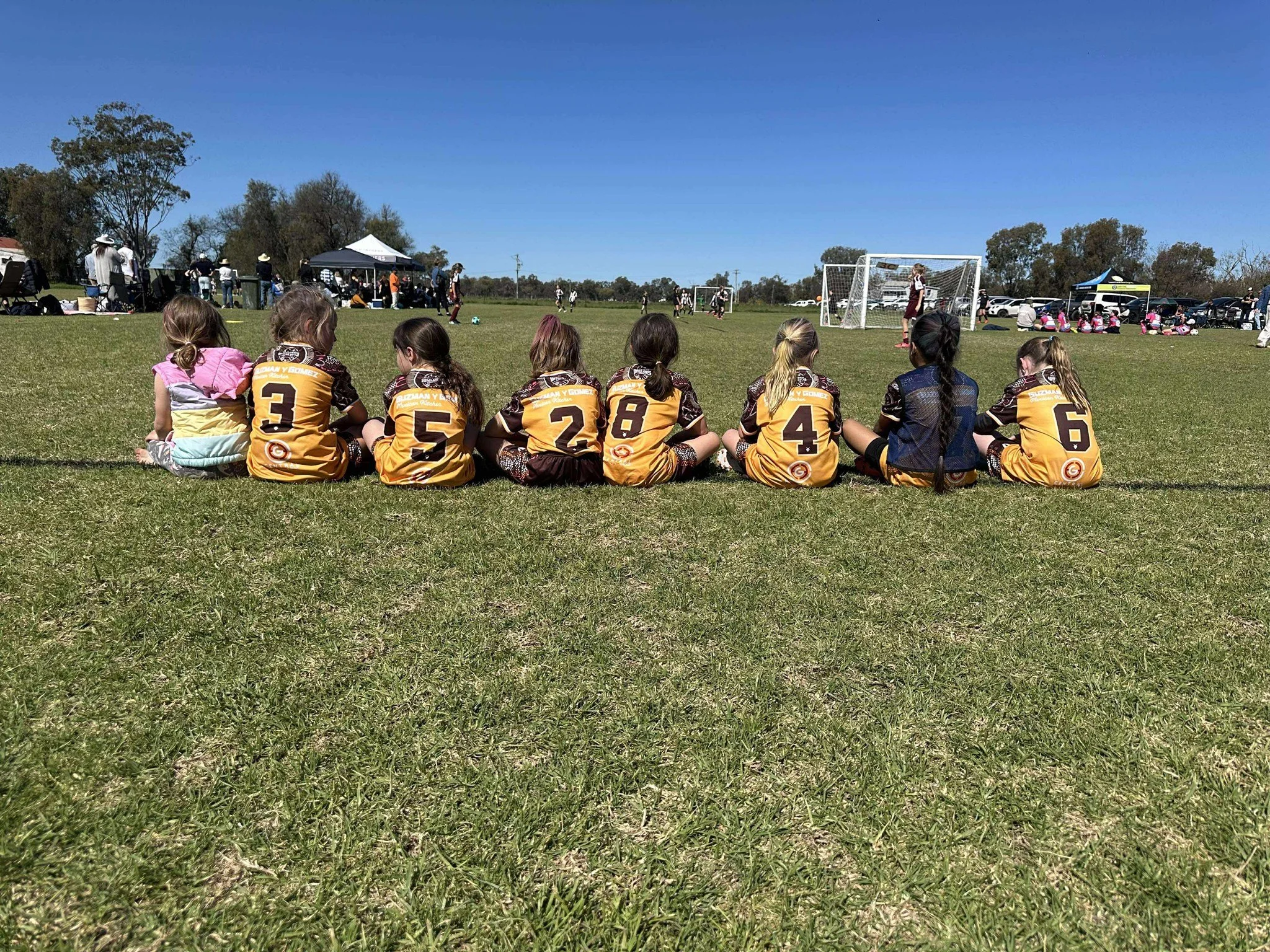 Group of young girls sitting on grass at a soccer game, wearing matching yellow and black jerseys, with a few spectators and players visible in the background under a clear blue sky.