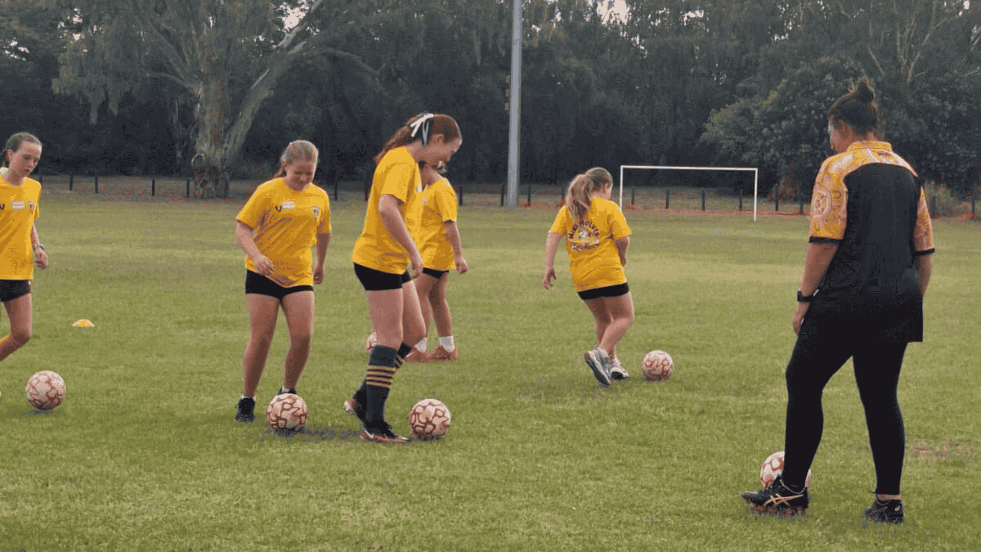 Young girls in yellow soccer uniforms practicing dribbling with soccer balls on a field, supervised by an adult coach.