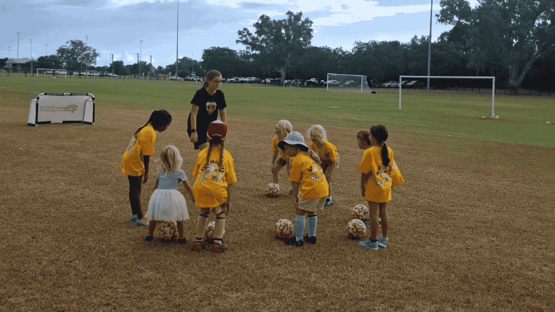 Children in yellow sports jerseys gather around soccer balls on a field during practice or game, with a coach supervising.