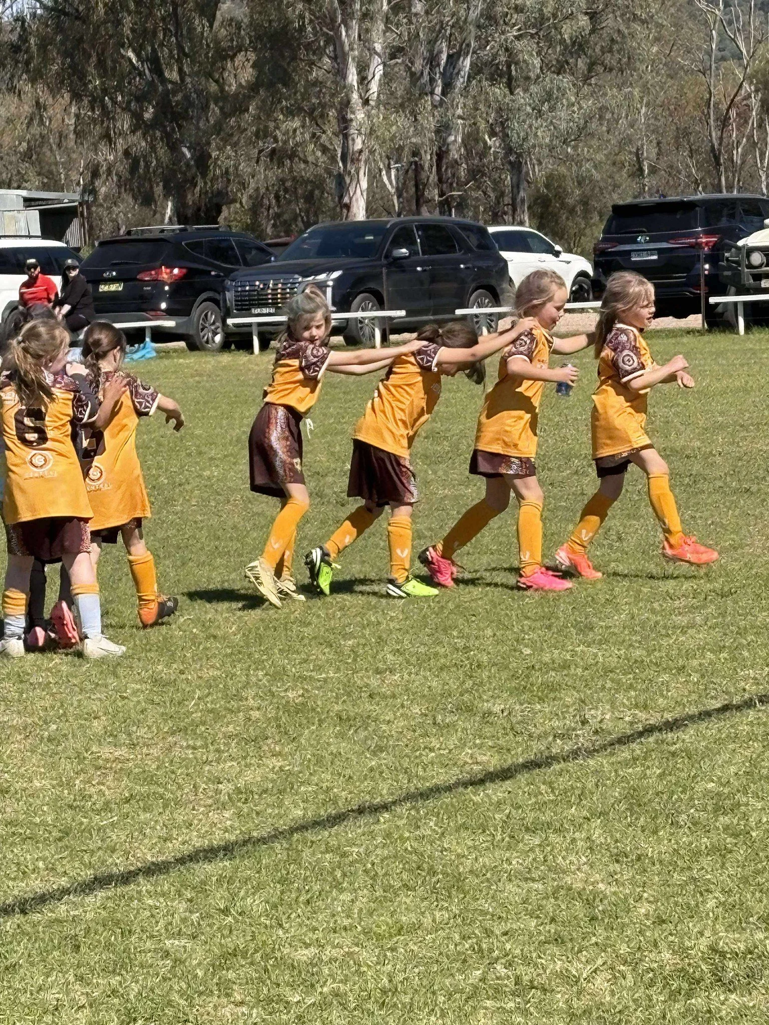 A group of young girls in orange and brown sports uniforms playing a game on a grassy field, holding onto each other in a line, with parked cars and trees in the background.