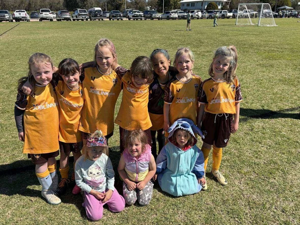 A group of young girls, some in sports uniforms and some in costumes, are posing on a grassy field after a soccer game. There are cars parked in the background and soccer goals on the field.