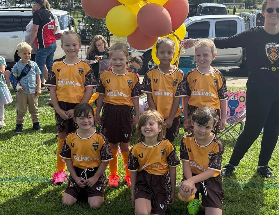 A group of young girls in soccer uniforms, five standing and three kneeling, posing outdoors with balloons in the background. The girls are smiling and have brown and blonde hair, some with ponytails. An adult woman stands to the right, smiling, with