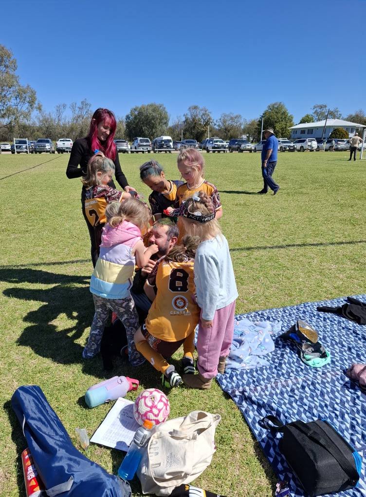 A group of children and a young woman are gathered on a grassy field, with children wearing sports jerseys, some with colorful hair. They appear to be playing or engaging in activities, with belongings and mats on the ground nearby. In the background