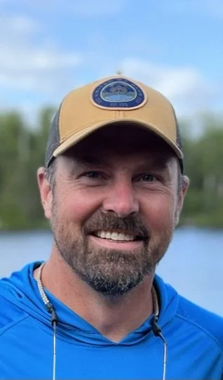 A man smiling outdoors, wearing a tan and gray baseball cap and a blue hoodie, with a body of water and trees in the background.
