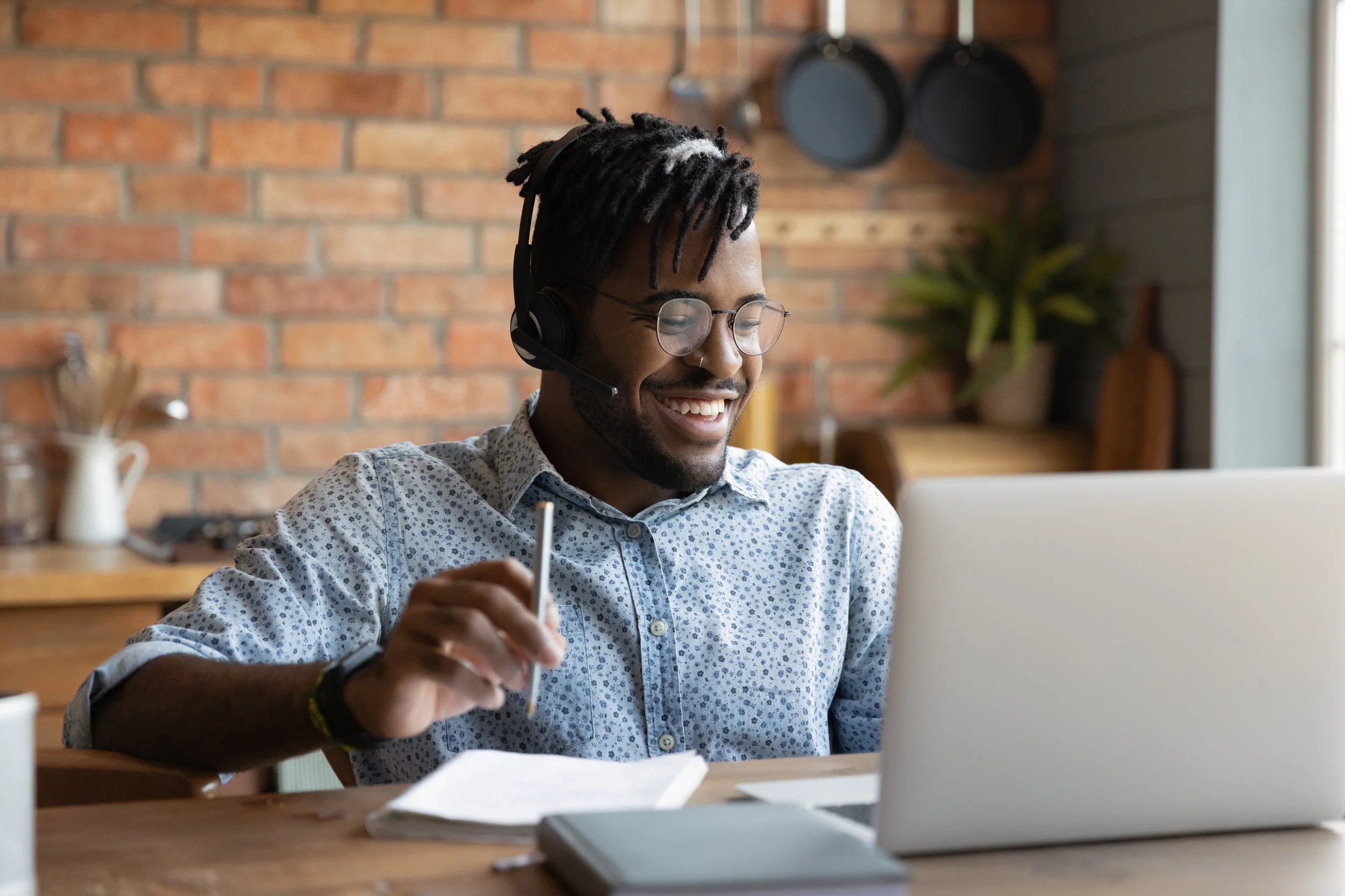 Man with glasses, wearing a headset, smiling while working on a laptop at a desk in a cozy kitchen with brick walls.