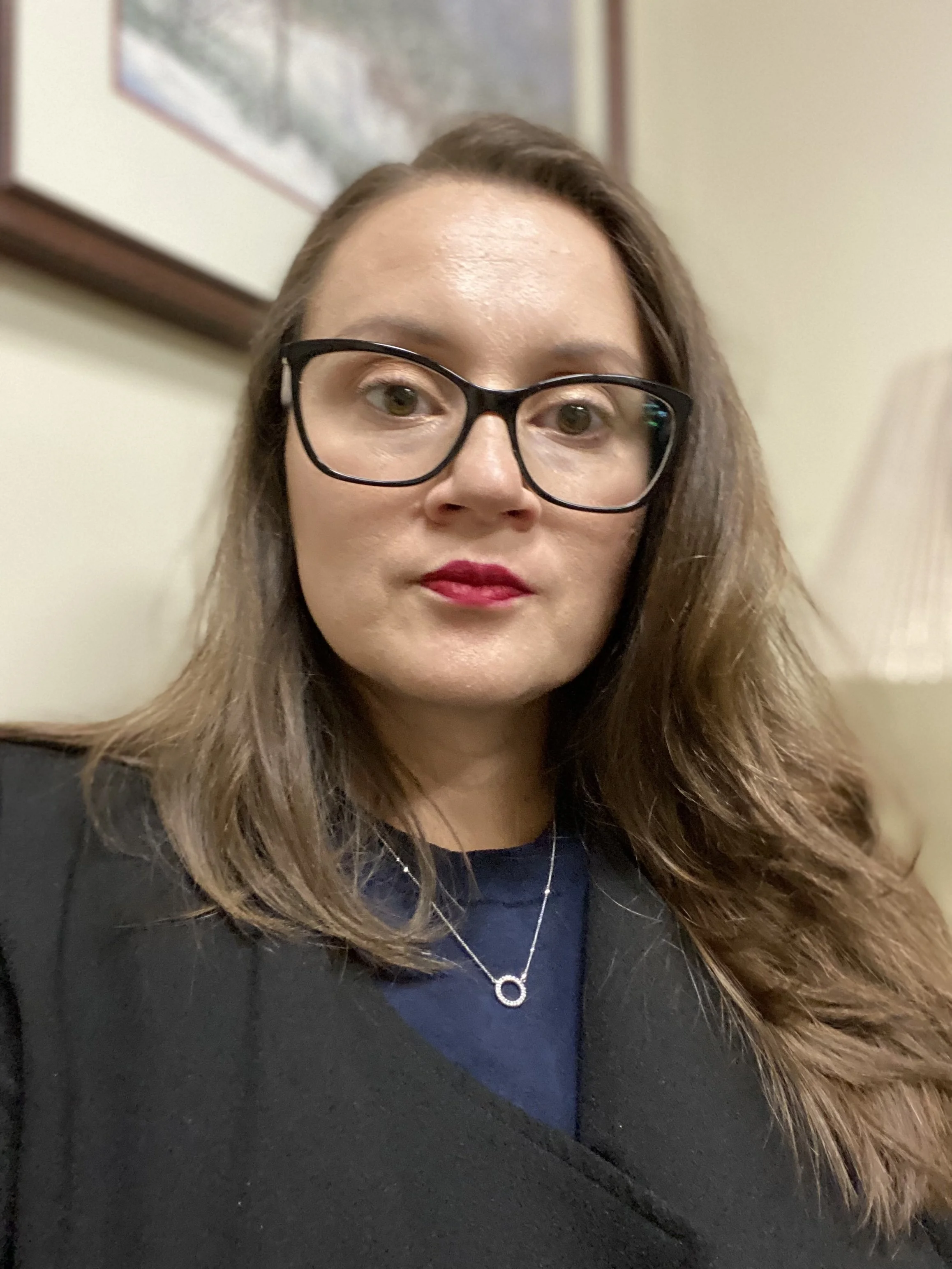 A woman with long brown hair, wearing black glasses, red lipstick, a black blazer over a dark blue top, and a silver necklace, taking a selfie indoors with a framed picture and a lamp in the background.