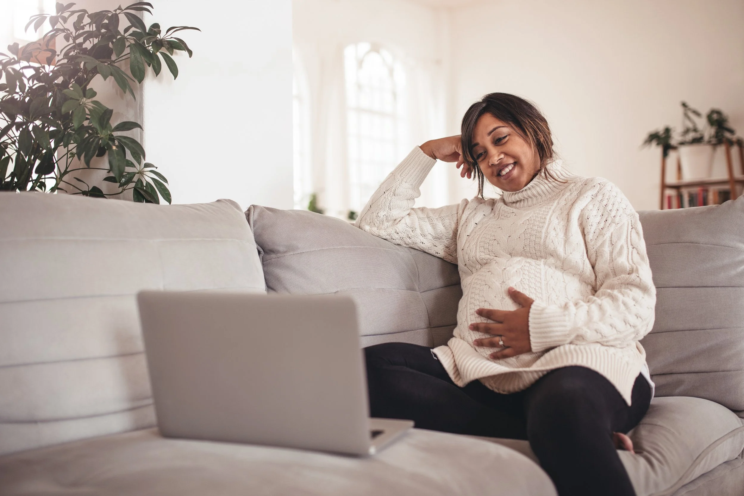 A pregnant woman sitting on a light-colored sofa, holding her belly and smiling, looking at a laptop on the couch. The room appears bright with natural light, decorated with houseplants and books.