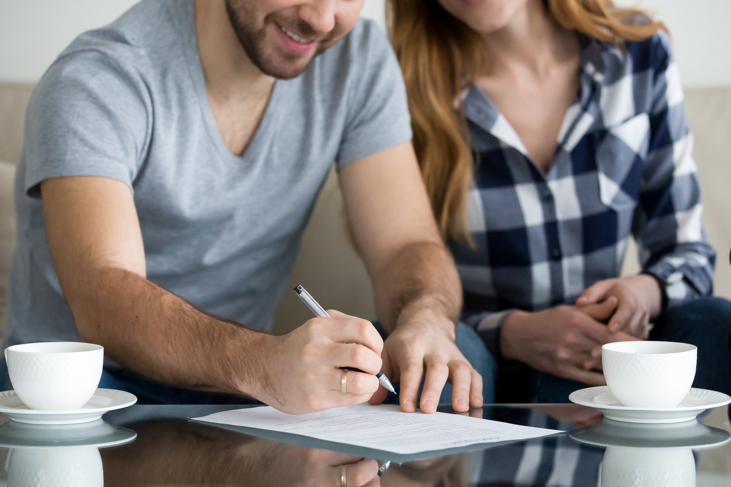 A man is signing a document on a table next to a woman, with two cups of tea or coffee nearby.