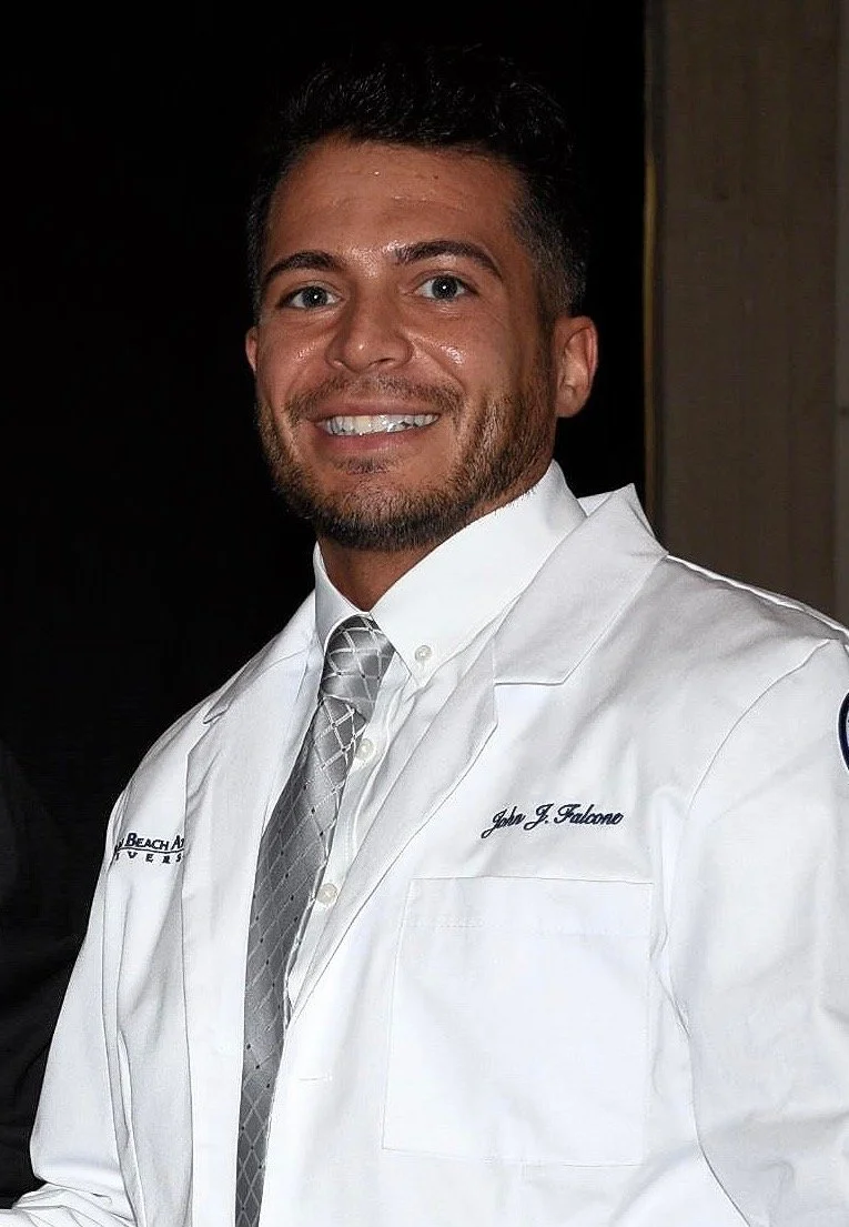 A man smiling and wearing a white coat with a name tag that says 'John J. Falcone' and a gray patterned tie.