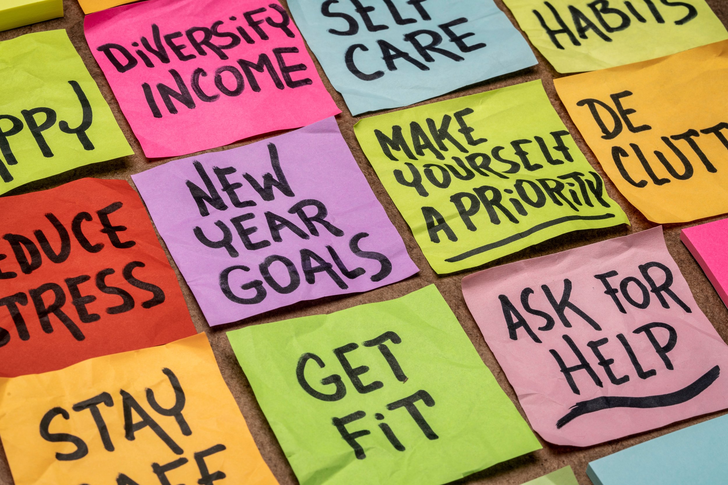Close-up of colorfully handwritten sticky notes with various motivational and health-related messages, including 'Diversify Income,' 'Self Care,' 'Habits,' 'Reduce Stress,' 'New Year Goals,' 'Make Yourself a Priority,' 'Get Fit,' and 'Ask for Help.'