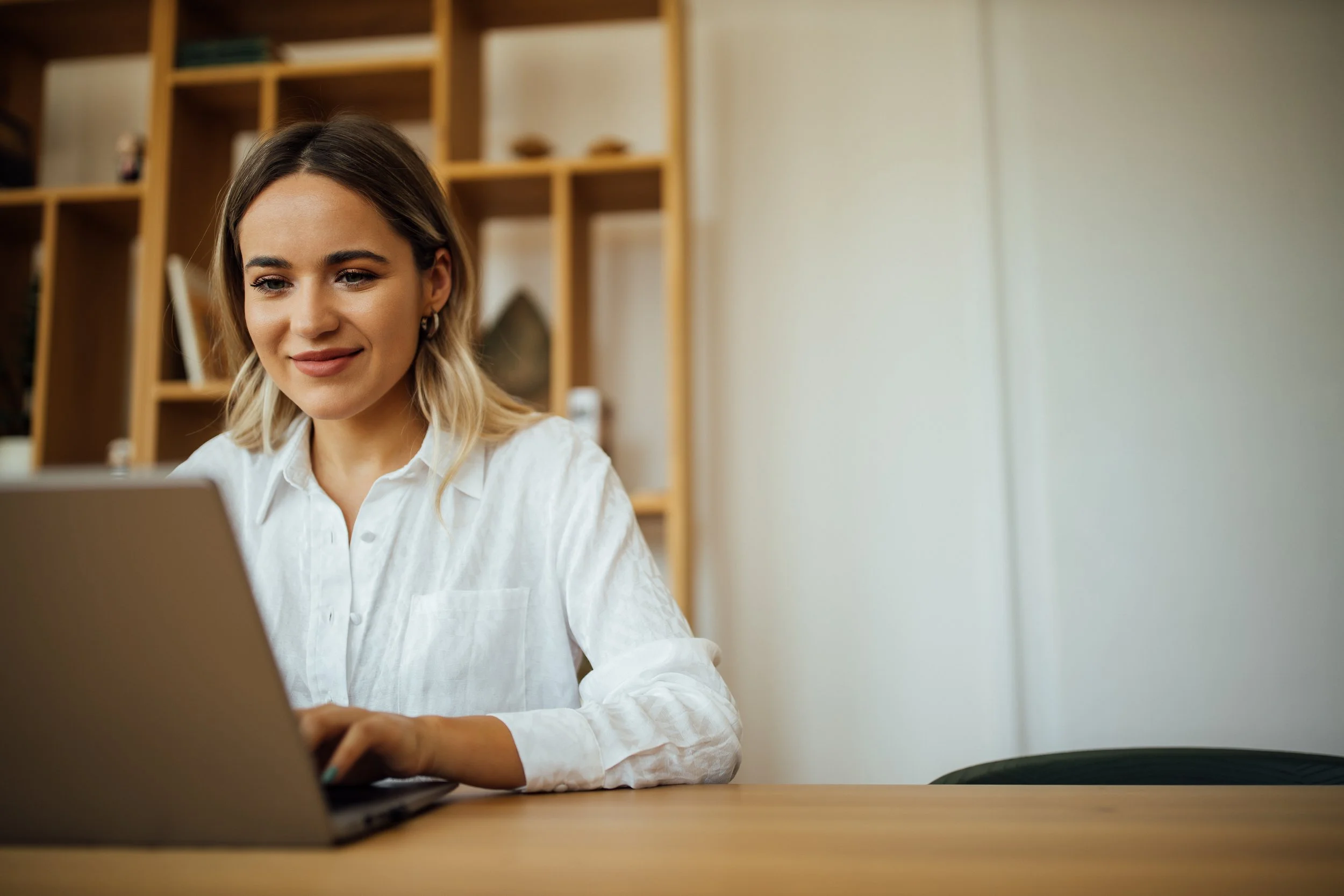 Young woman with shoulder-length blonde hair working on a laptop at a wooden table, with wooden shelves in the background.