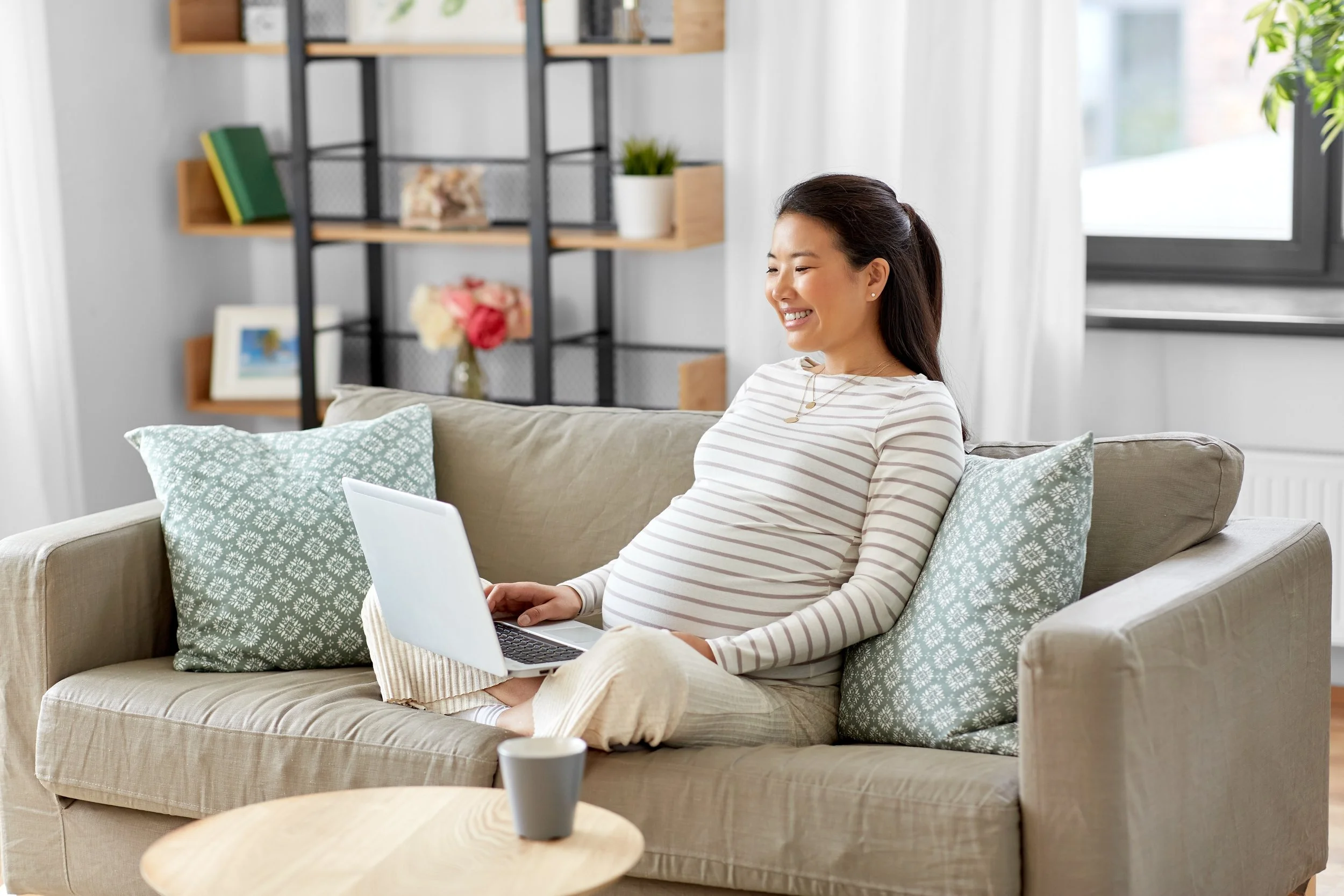 A pregnant woman sitting on a beige sofa in a bright living room, using a laptop, with decorative pillows, a coffee cup on a small wooden table, and a bookshelf with plants and framed photos in the background.
