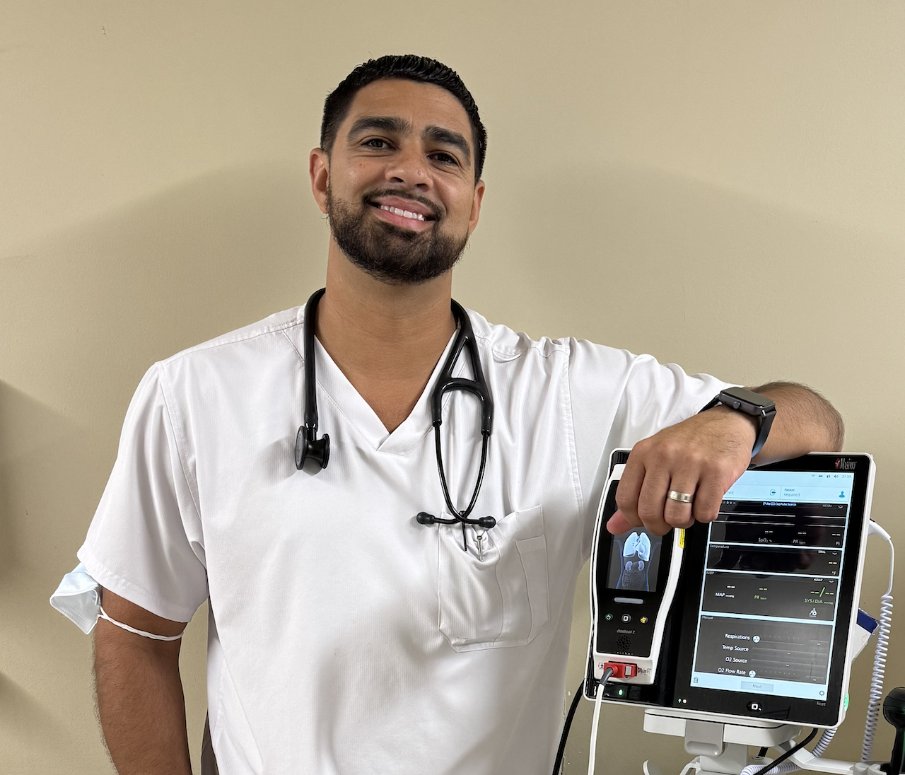 A smiling male healthcare professional wearing white scrubs and a stethoscope around his neck, standing next to a medical monitor in a clinical setting.