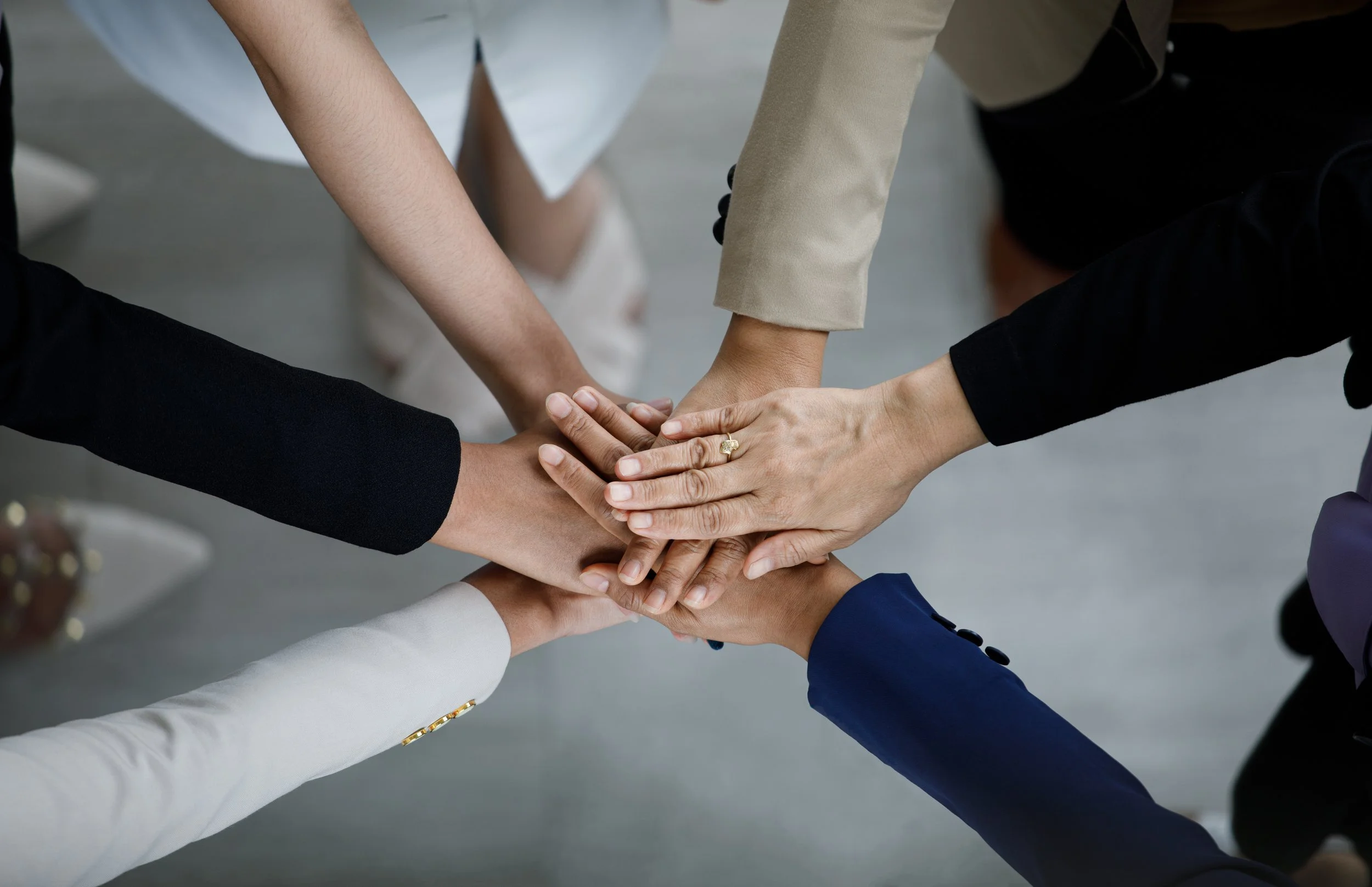 Six people in business attire placing their hands together in a show of teamwork and unity.